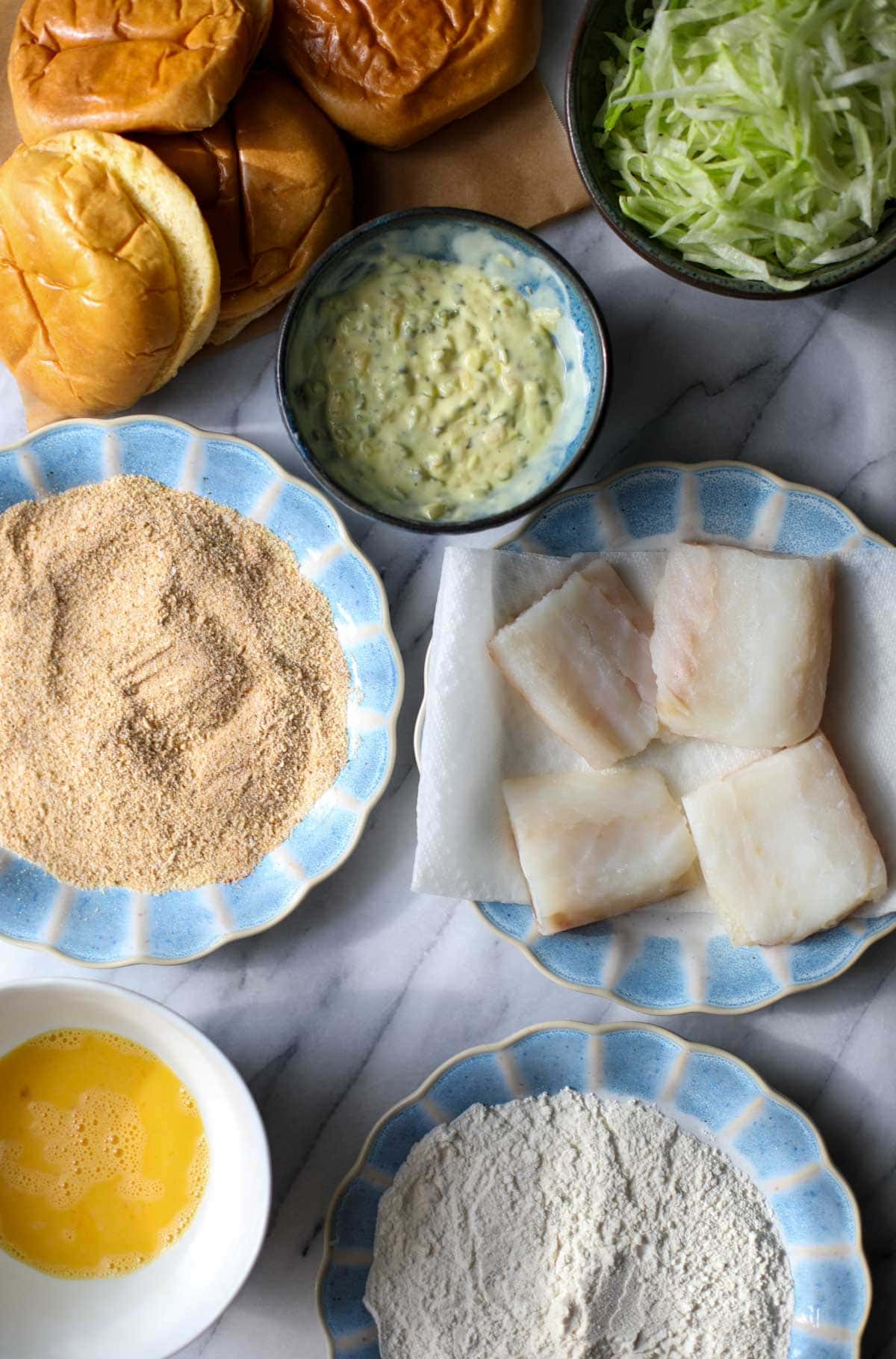 The ingredients to make a fried fish sandwich including blue rimmed plates with breadcrumbs, cod filets, and flour, a blue bowl of tartar sauce, several fresh brioche buns, shredded iceberg lettuce, and a white bowl with a scrambled egg.