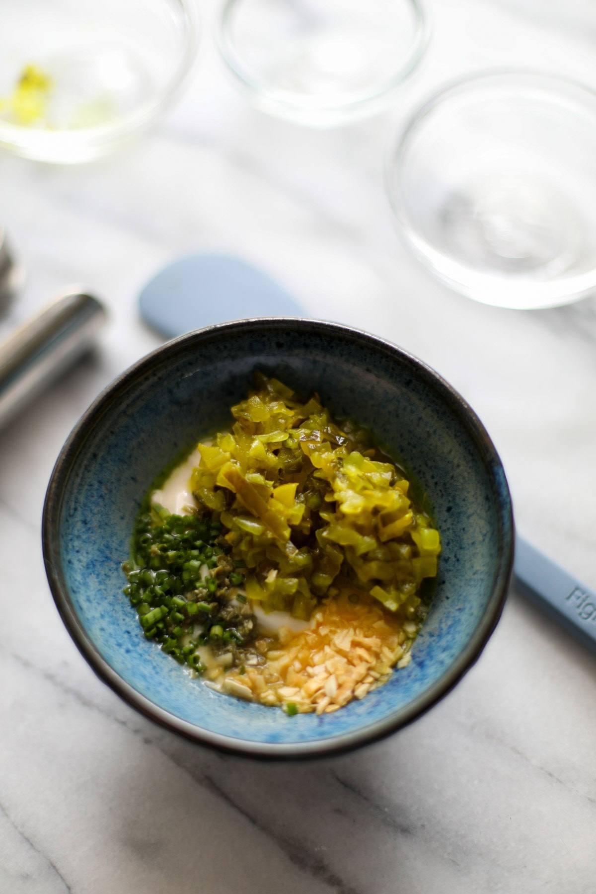 A small blue bowl filled with homemade tartar sauce topped with chives and extra dill relish with a lemon, jar of vlasic dill pickle relish, and capers in the background.
