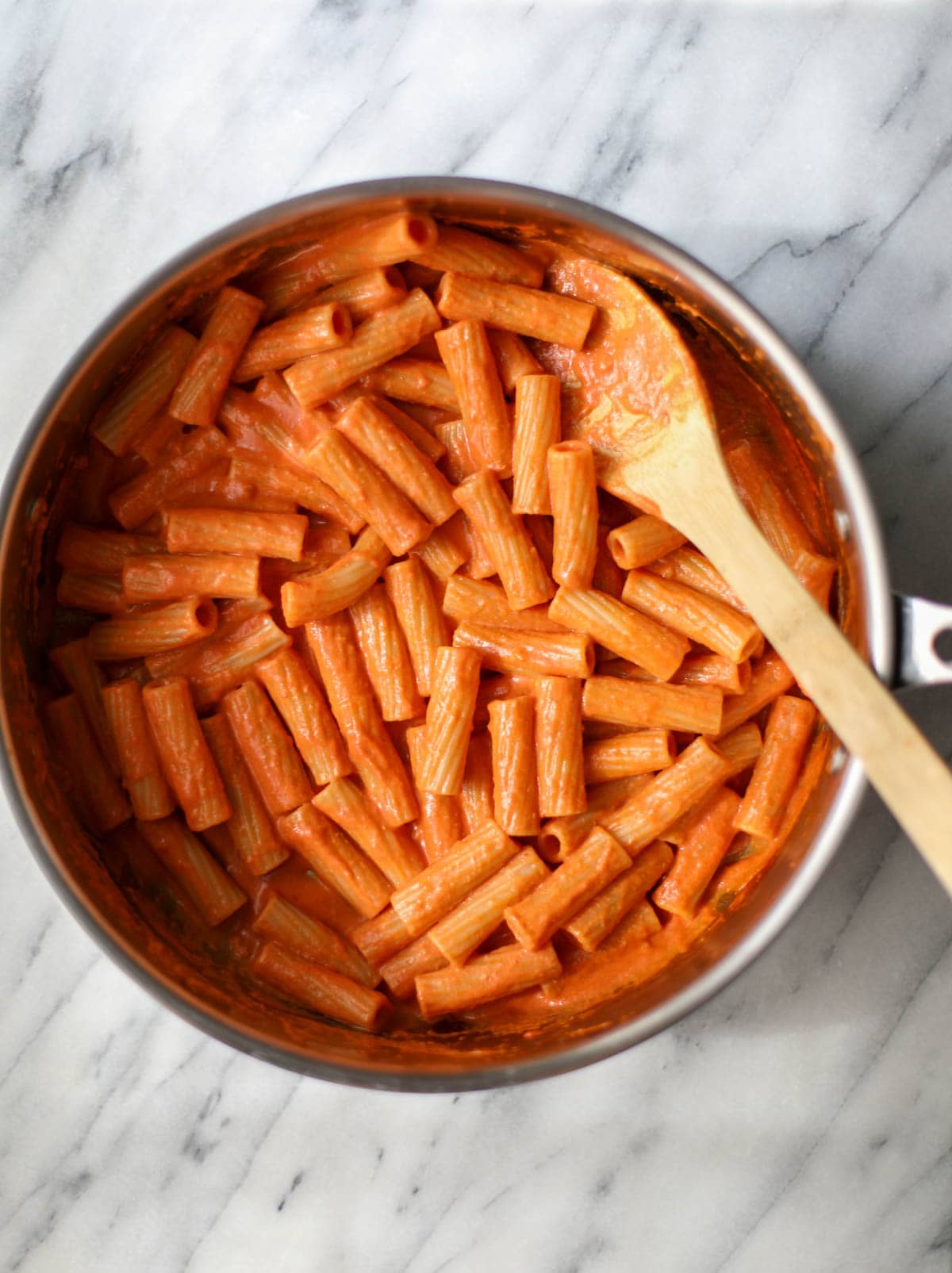 A large stainless steel skillet filled with orangey red vodka sauce pasta with a wooden spoon hanging off the edge.