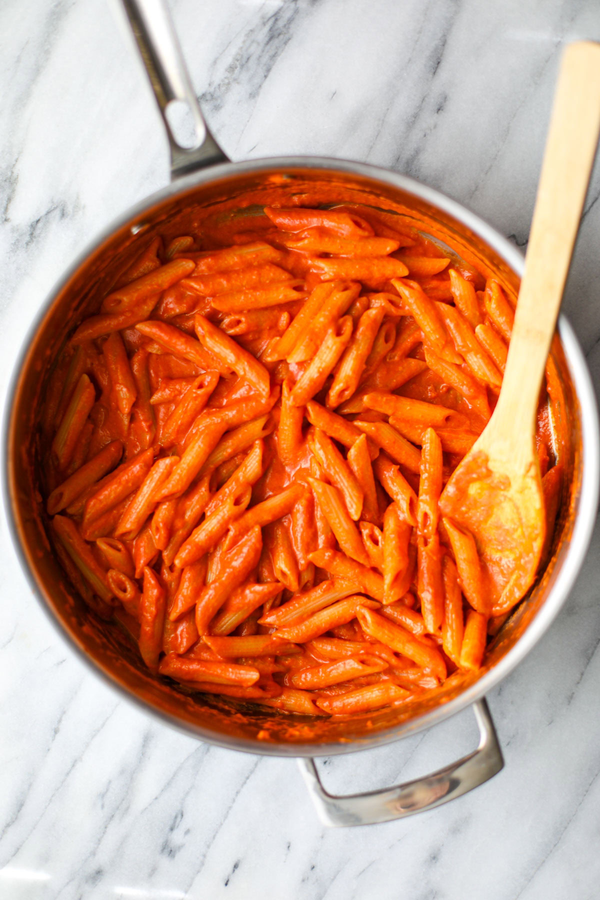 A large stainless steel skillet filled with orangey red vodka sauce pasta with a wooden spoon hanging off the edge.