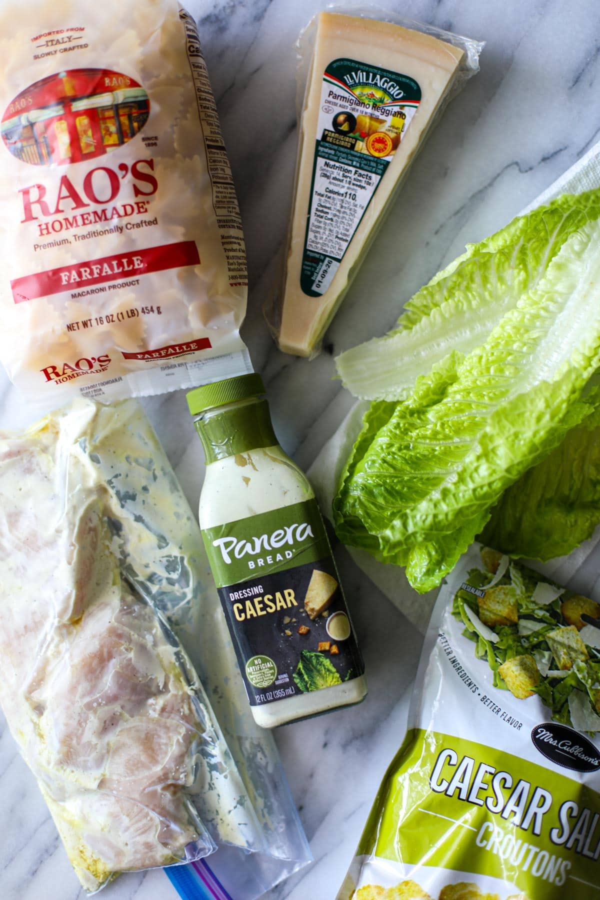 All the ingredients laid out on a white marble counter to make chicken caesar pasta salad including chicken breasts, a bag of croutons, a head of romaine lettuce, a glass jar of caesar dressing, a bag of farfalle pasta and a wedge of parmigiano reggiano.