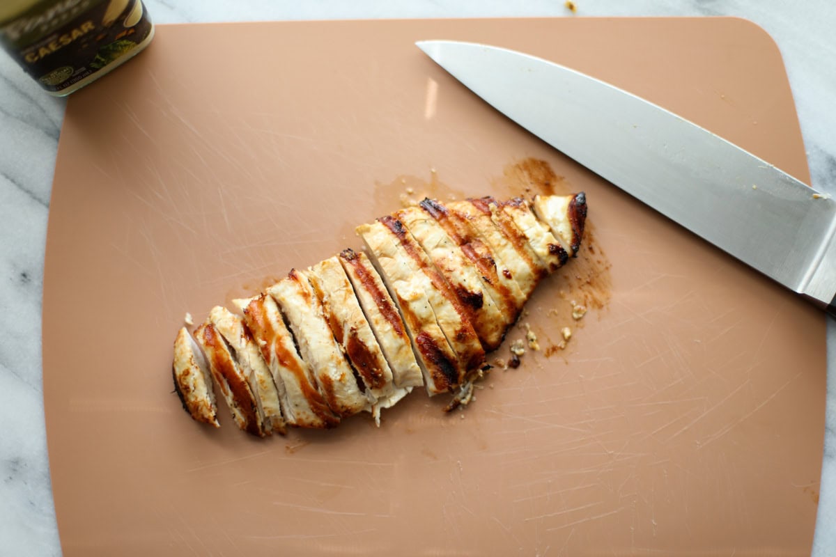 Grilled chicken cut into slices on a terracotta cutting board with a chef's knife set next to it. 