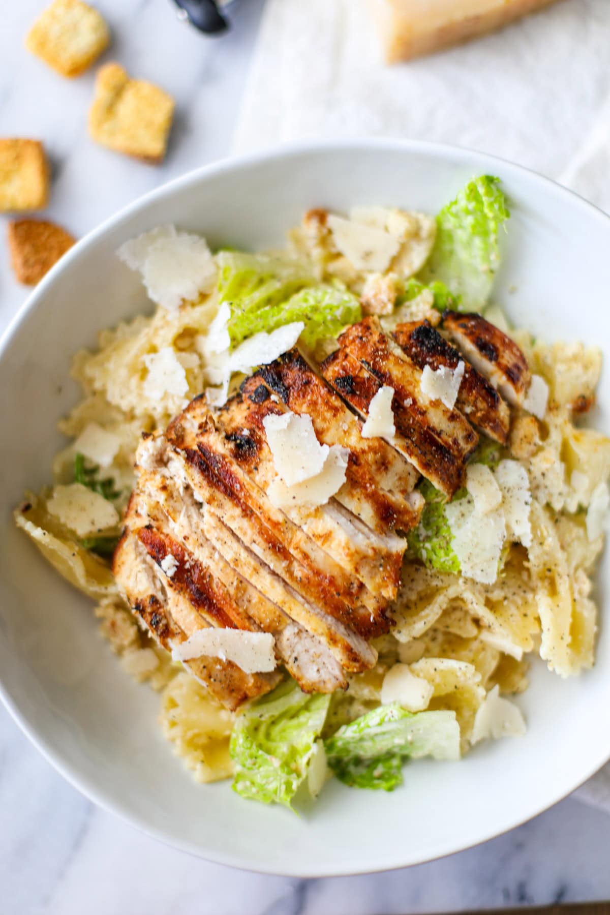 Large white shallow bowl filled with romaine lettuce, farfalle pasta, grilled chicken, and shaved parmigiano reggiano with croutons, a wedge of parmigiano reggiano, and a cheese slicer in the background. 