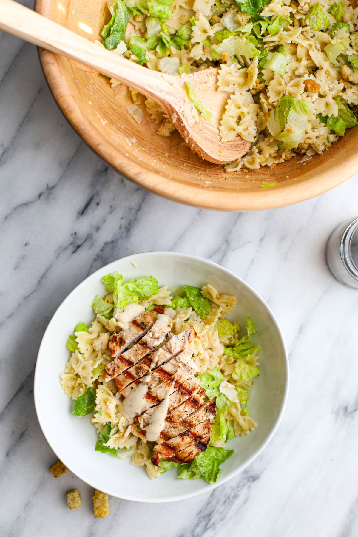 Large white shallow bowl filled with romaine lettuce, farfalle pasta, grilled chicken, and shaved parmigiano reggiano with croutons, and a wood bowl in the background.