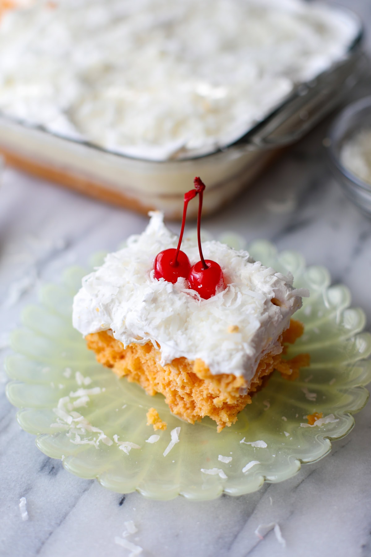 A slice of Pina colada cake with a cool whip topping and sweetened coconut flakes with maraschino cherries on top resting on a light yellow, scalloped edge plate with sweetened coconut crumbs sprinkled around.
