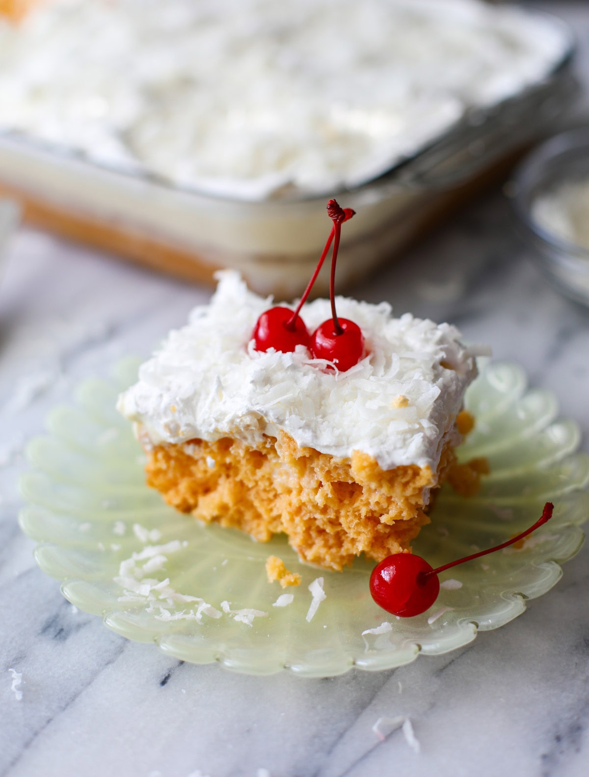 A slice of Pina colada cake with a cool whip topping and sweetened coconut flakes with maraschino cherries on top resting on a light yellow, scalloped edge plate with sweetened coconut crumbs sprinkled around.
