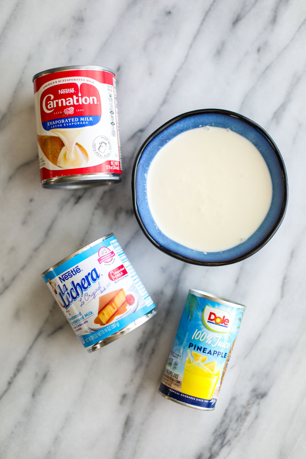 A can of evaporated milk, a bowl of heavy cream, a can of sweetened condensed milk, and a can of pineapple juice.