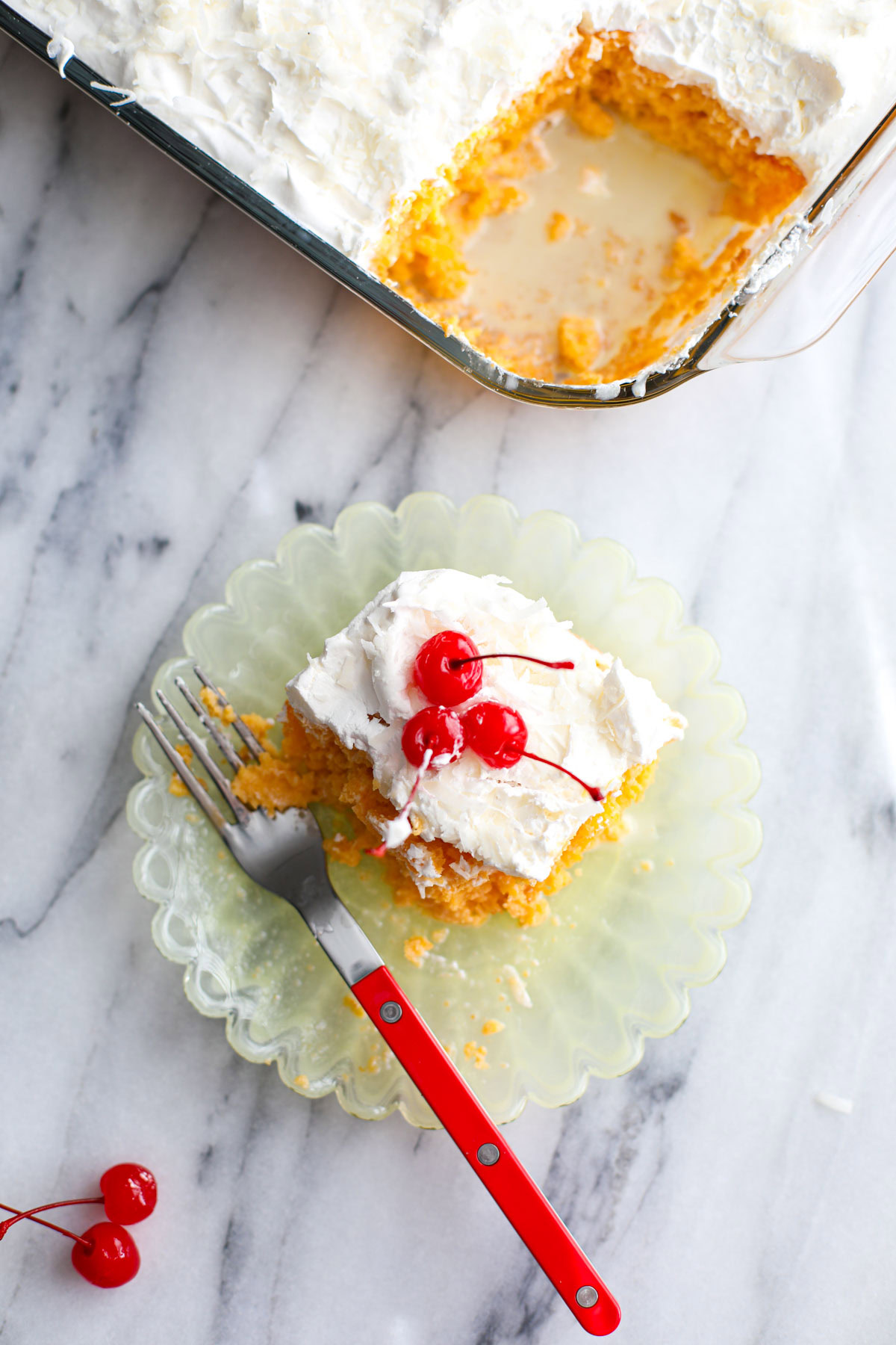 A piece of piña colada cake on a yellow plate topped with cherries with a red fork.