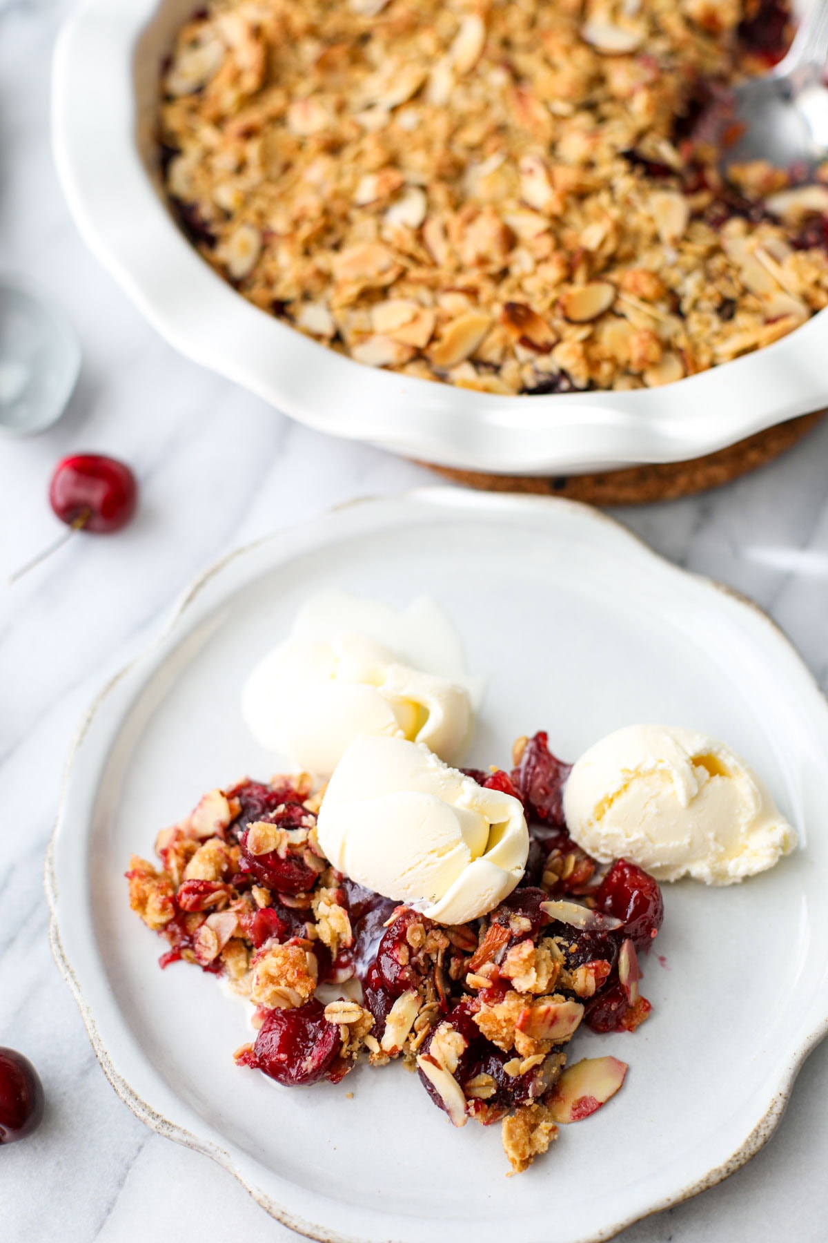 A ruffled and distressed white plate with a scoop of cherry crisp with golden brown topping and three small scoops of vanilla ice cream.