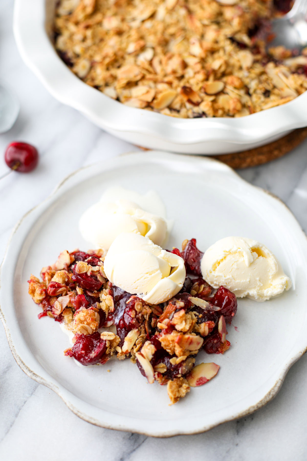 A ruffled and distressed white pie plate with a scoop of cherry crisp with golden brown topping and three small scoops of vanilla ice cream.