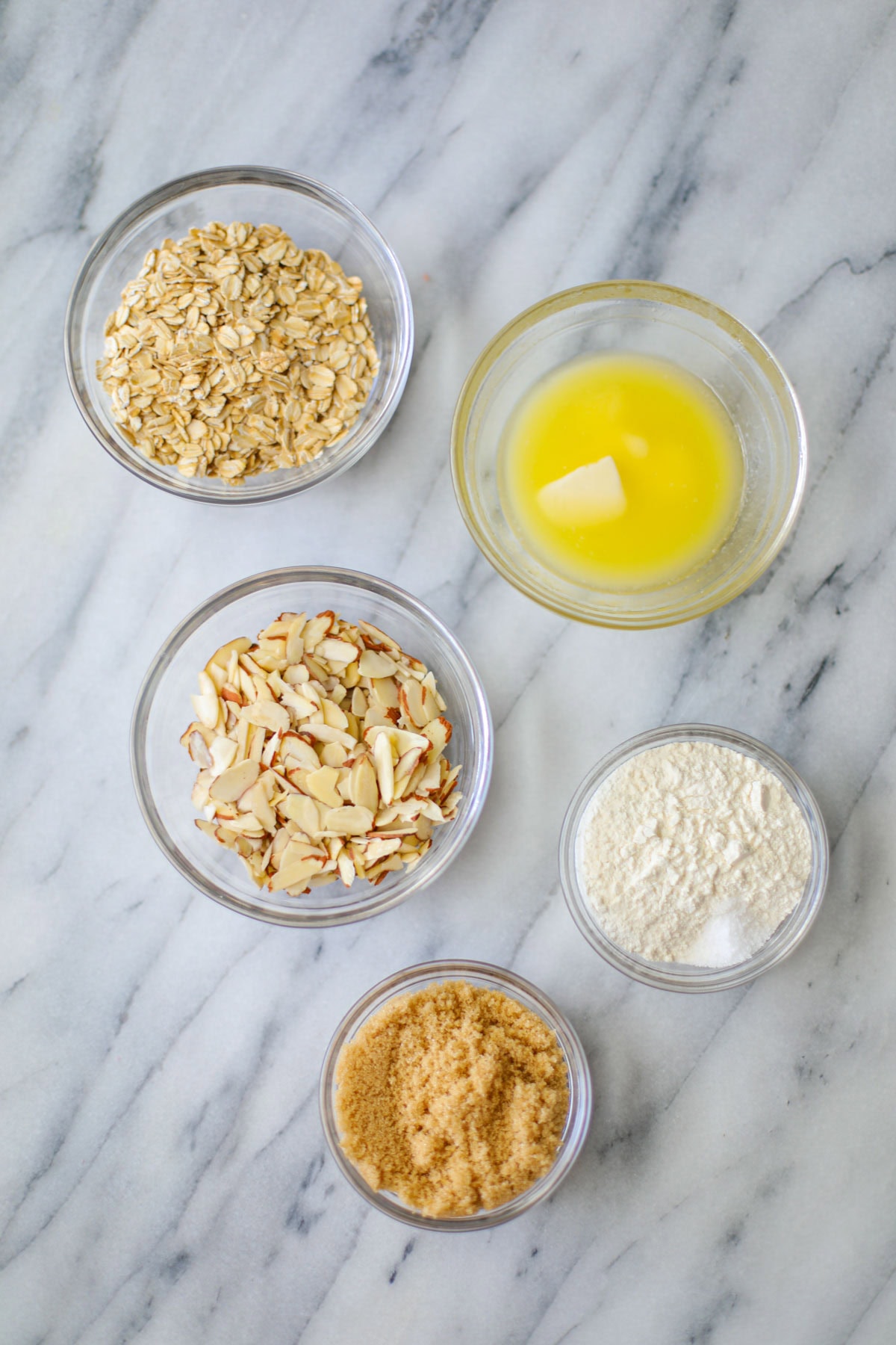 Five glass prep bowls, filled with brown sugar, flour, sliced almonds, and melted butter atop a white marble board.