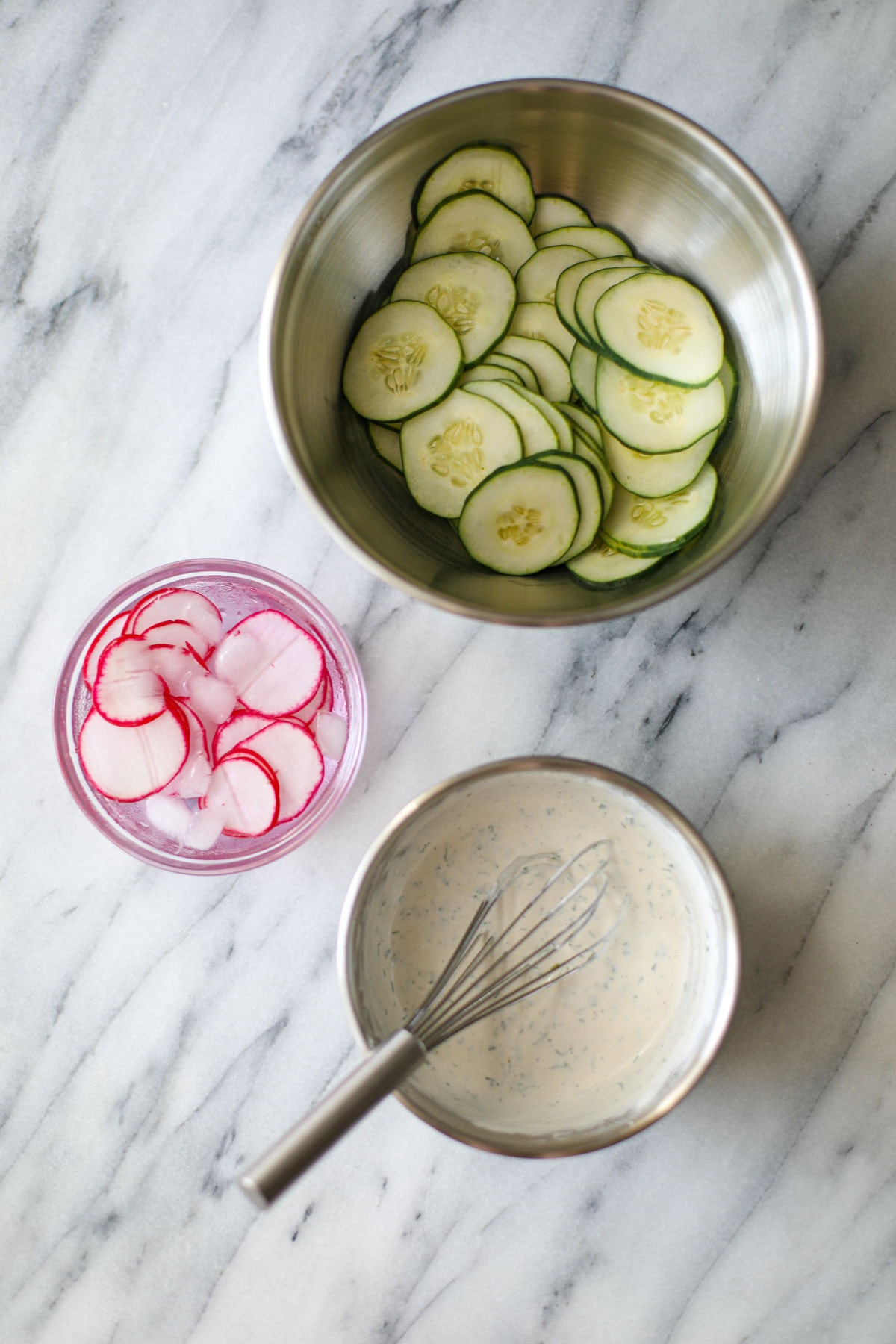 A stainless steel mixing bowl of sliced cucumbers, a small glass bowl with radishes sitting in ice water, and a smaller stainless steel mixing bowl with a sour cream dressing with a whisk hanging on the edge all seated on a marble counter.