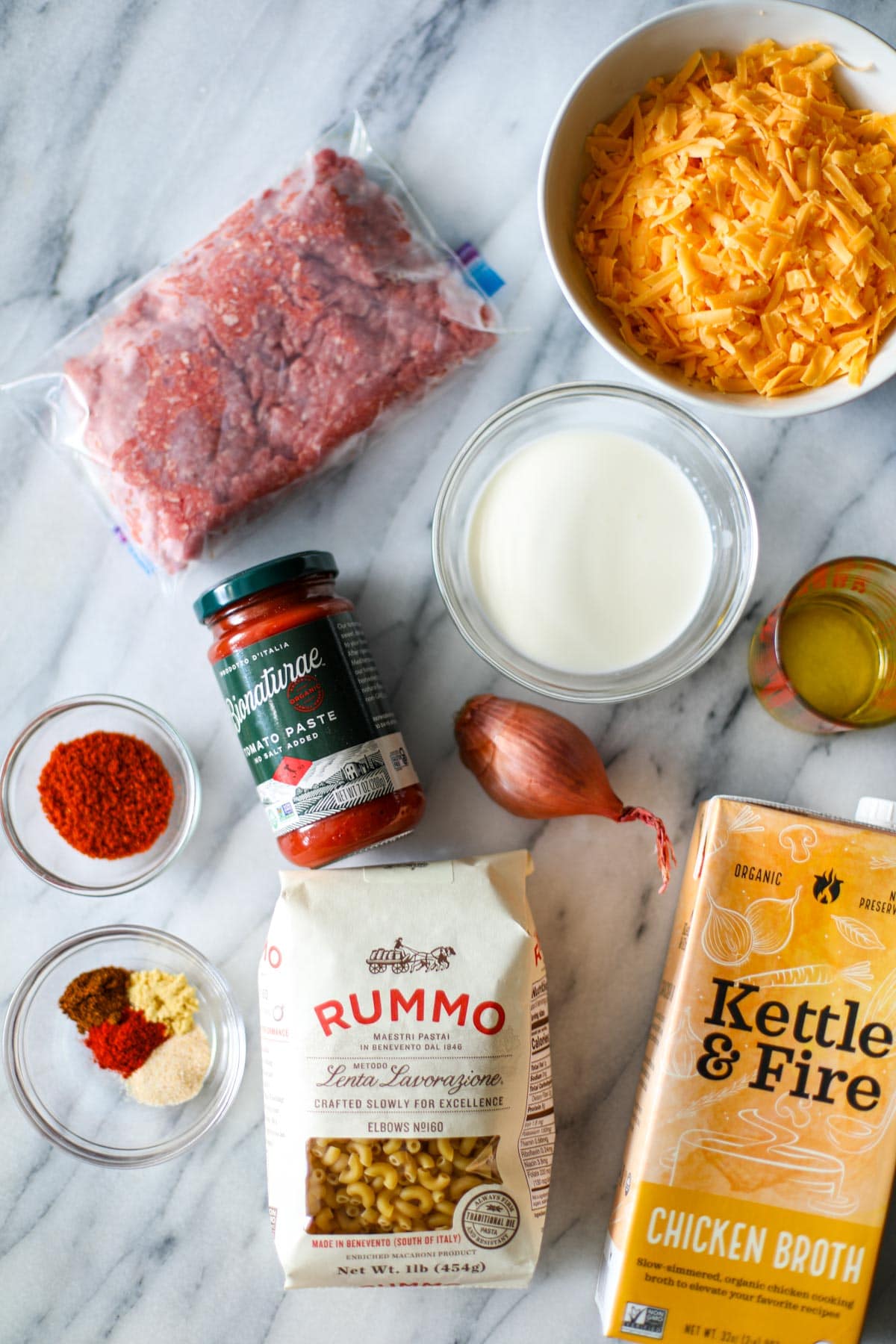 All the ingredients for hamburger helper laid out on a marble countertop including a package of ground sirloin, a white bowl filled with cheddar cheese, a small glass bowl with heavy cream, a measuring cup with olive oil, a jar of tomato paste, a shallot, two small glass bowls of seasoning, a package of elbow noodles, and a carton of chicken broth.