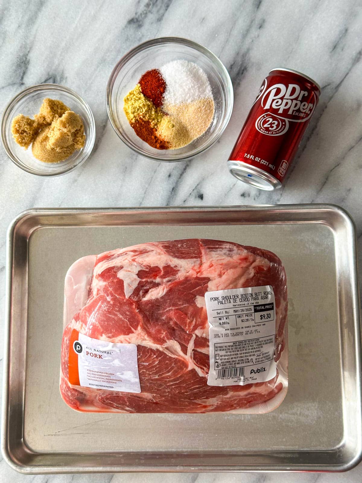 A glass bowl with brown sugar, another glass bowl filled with red, tan and white seasonings, a mini can of Dr. Pepper, and a packaged Boston butt roast sitting on a gold cookie sheet.
