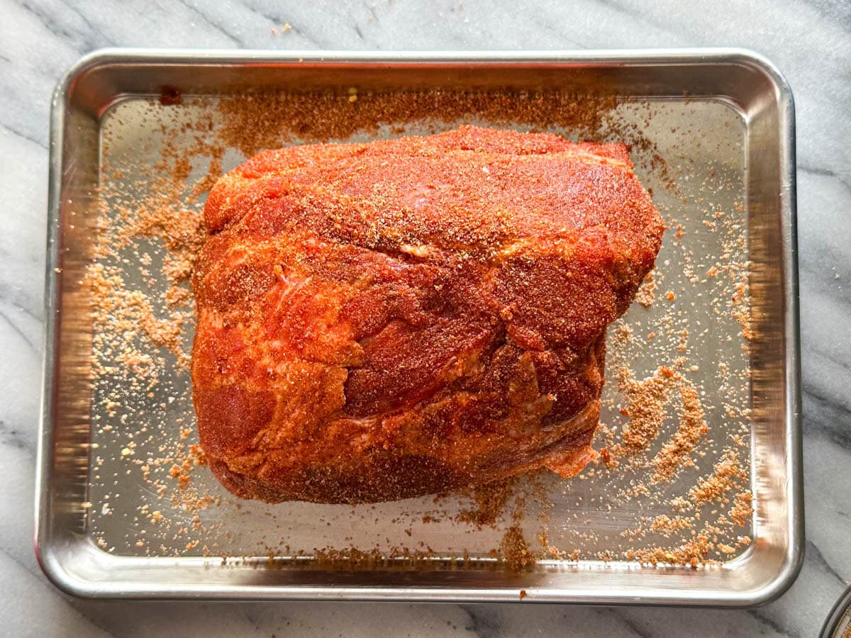 A Boston butt roast covered in a reddish orange seasoning blend sat on a metal baking pan.