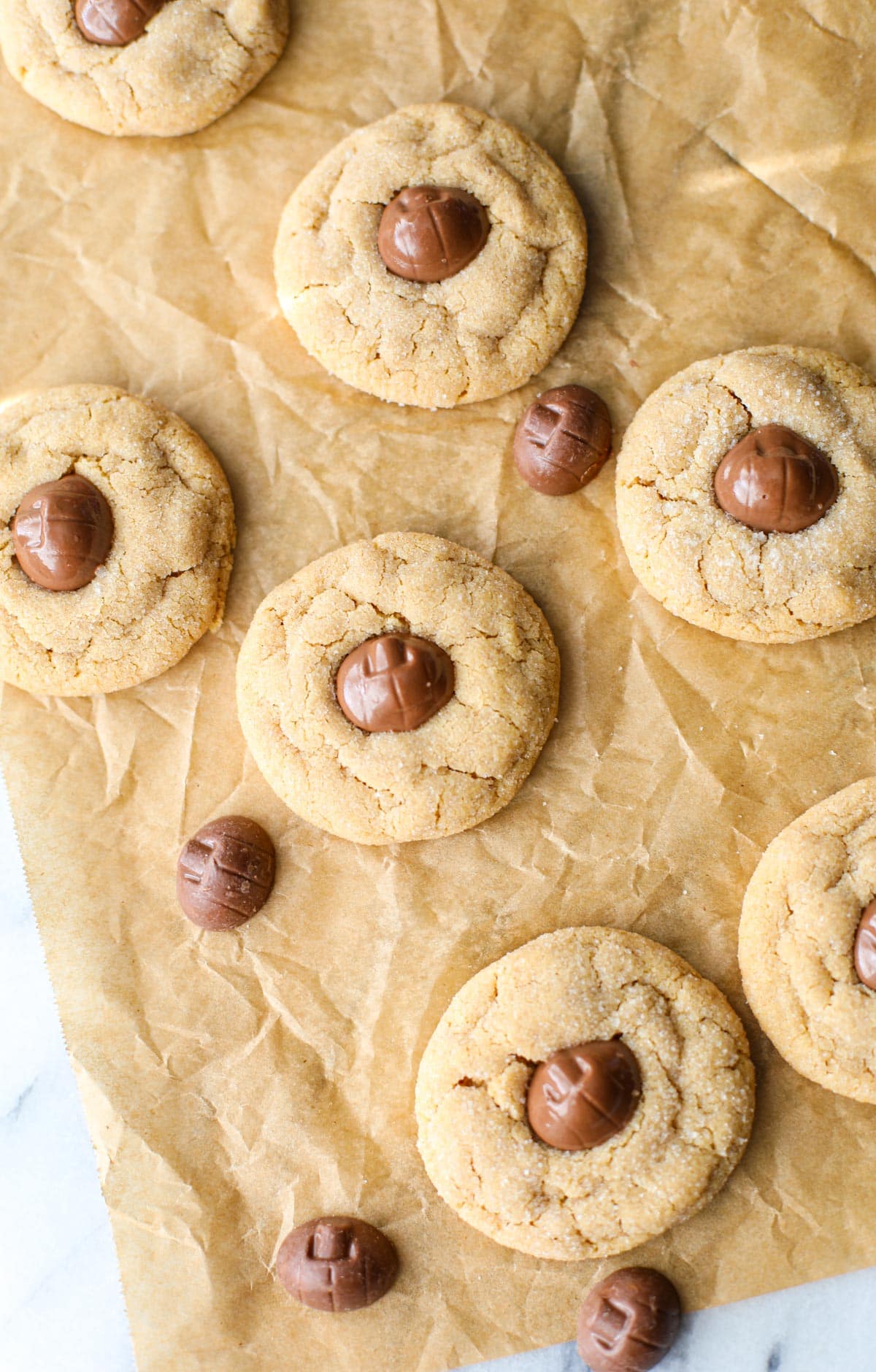 A crinkled brown piece of parchment paper with 6 Mini Reese's Pumpkin Peanut Butter Blossoms on top that have golden crackly edges and pumpkin shaped chocolates placed in the center. 