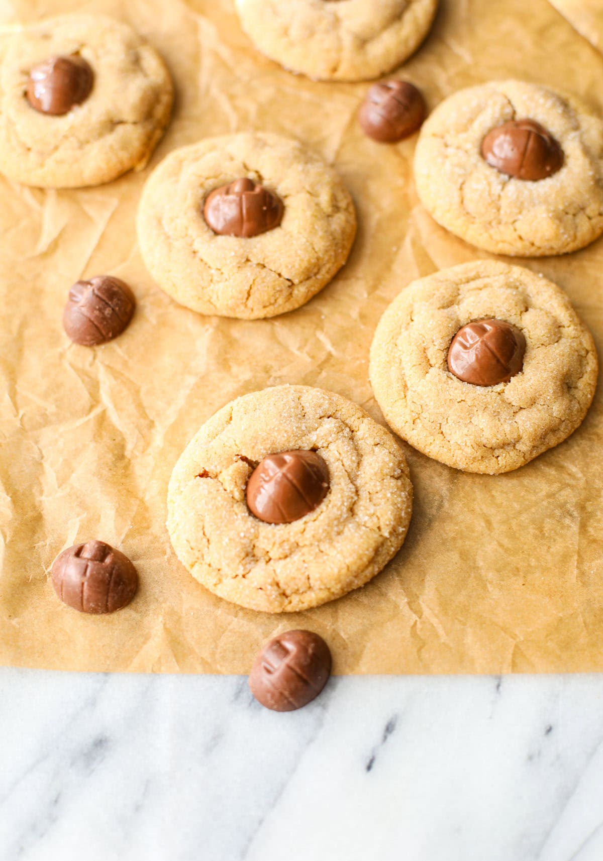 A crinkled brown piece of parchment paper with 6 Mini Reese's Pumpkin Peanut Butter Blossoms on top that have golden crackly edges and pumpkin shaped chocolates placed in the center. 