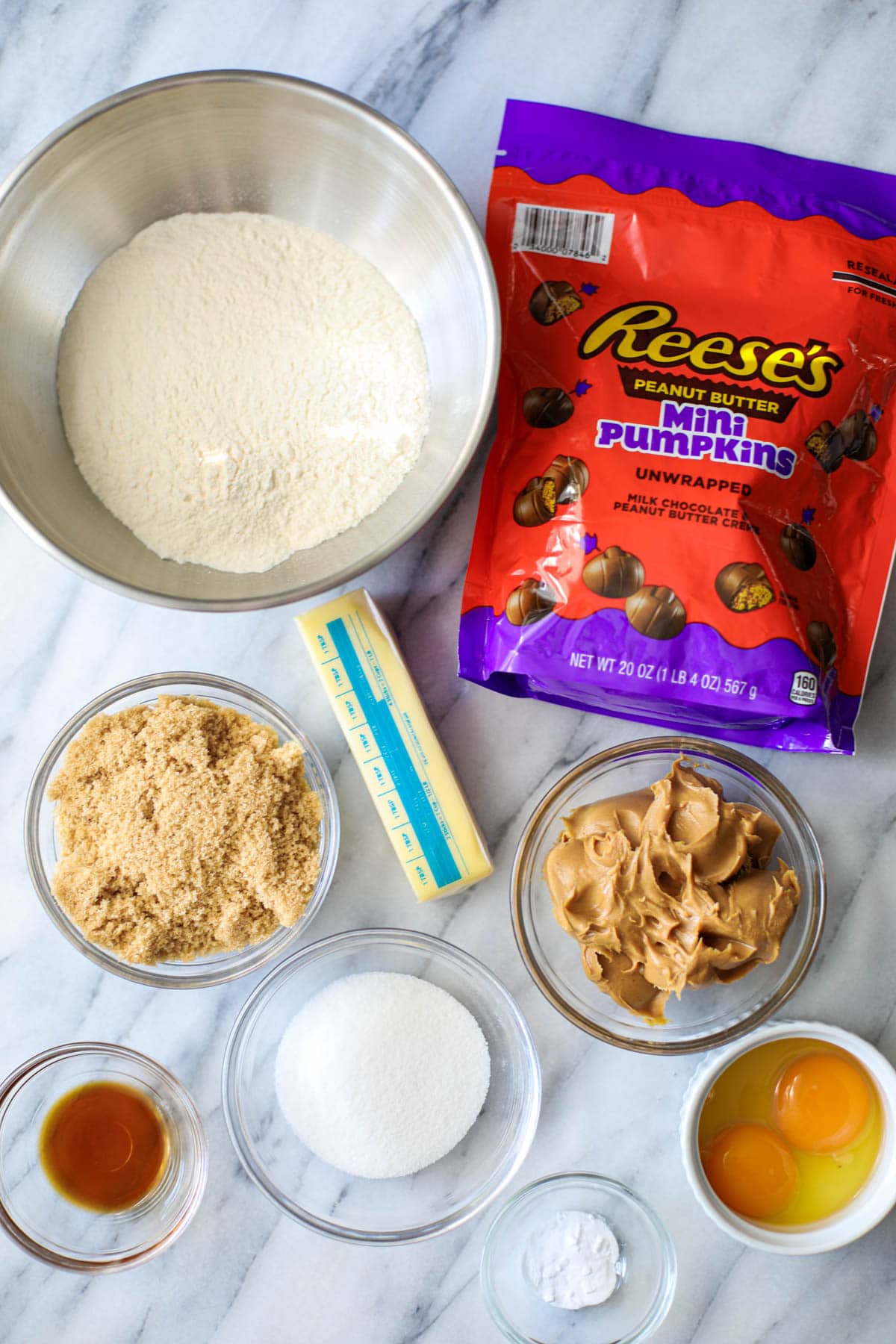 A stainless steel bowl filled with flour, a orange and purple package of Reese's peanut butter mini pumpkins, a wrapped stick of butter, a glass bowl of light brown sugar, a glass bowl of creamy peanut butter, a glass bowl of white granulated sugar, and three small bowls filled with vanilla, 2 eggs, and baking soda all set atop a marble countertop.