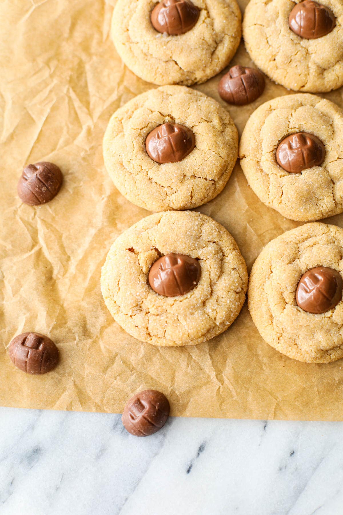 A crinkled brown piece of parchment paper with two rows of Mini Reese's Pumpkin Peanut Butter Blossoms on top that have golden crackly edges and pumpkin shaped chocolates placed in the center with several stray pumpkin chocolates scattered around.