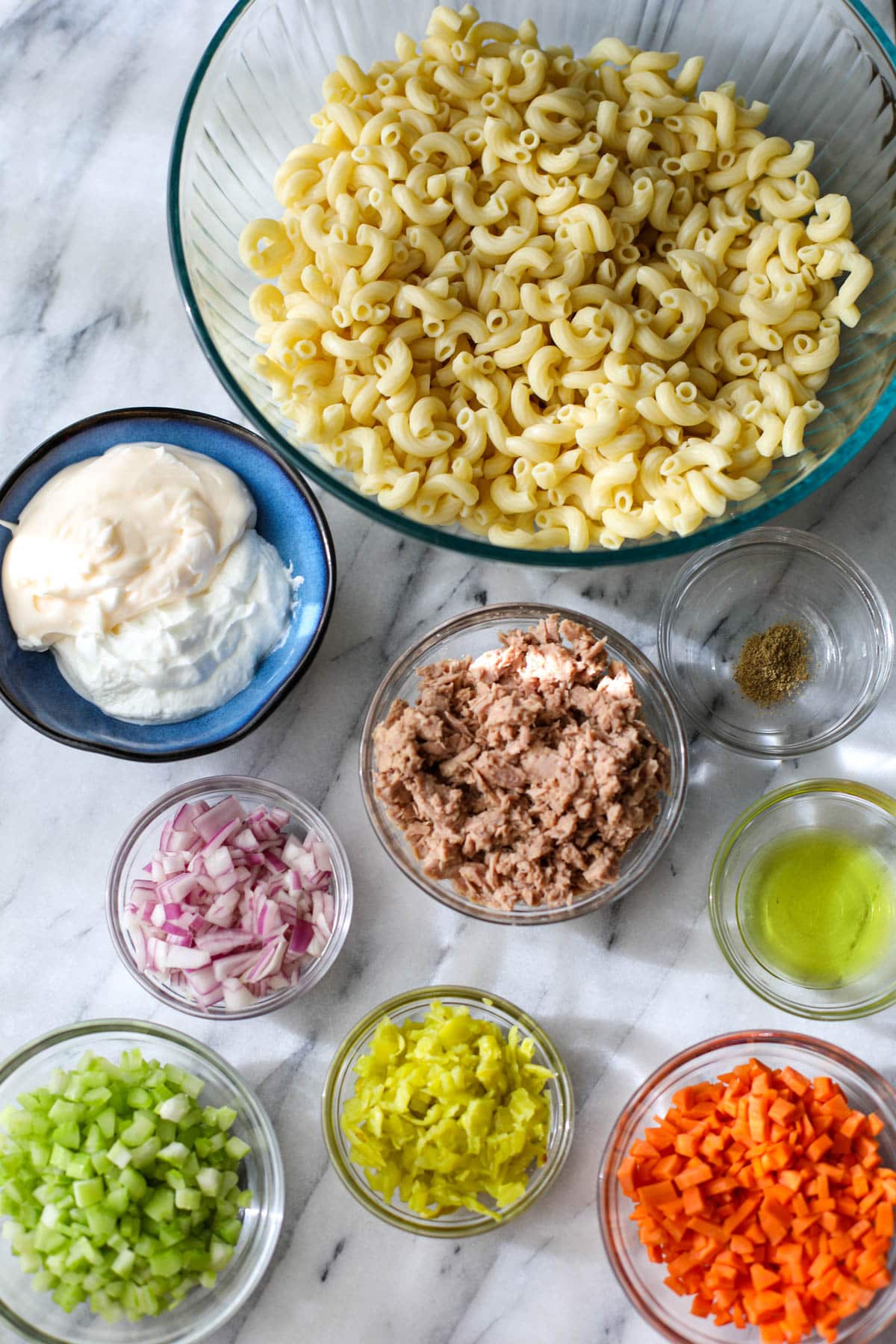Ingredients laid out on a marble table including a blue bowl with mayonnaise and yogurt, a clear bowl with elbow pasta, and various small glass bowls with tuna, red onion, pepperoncinis, celery, carrots, and celery salt.