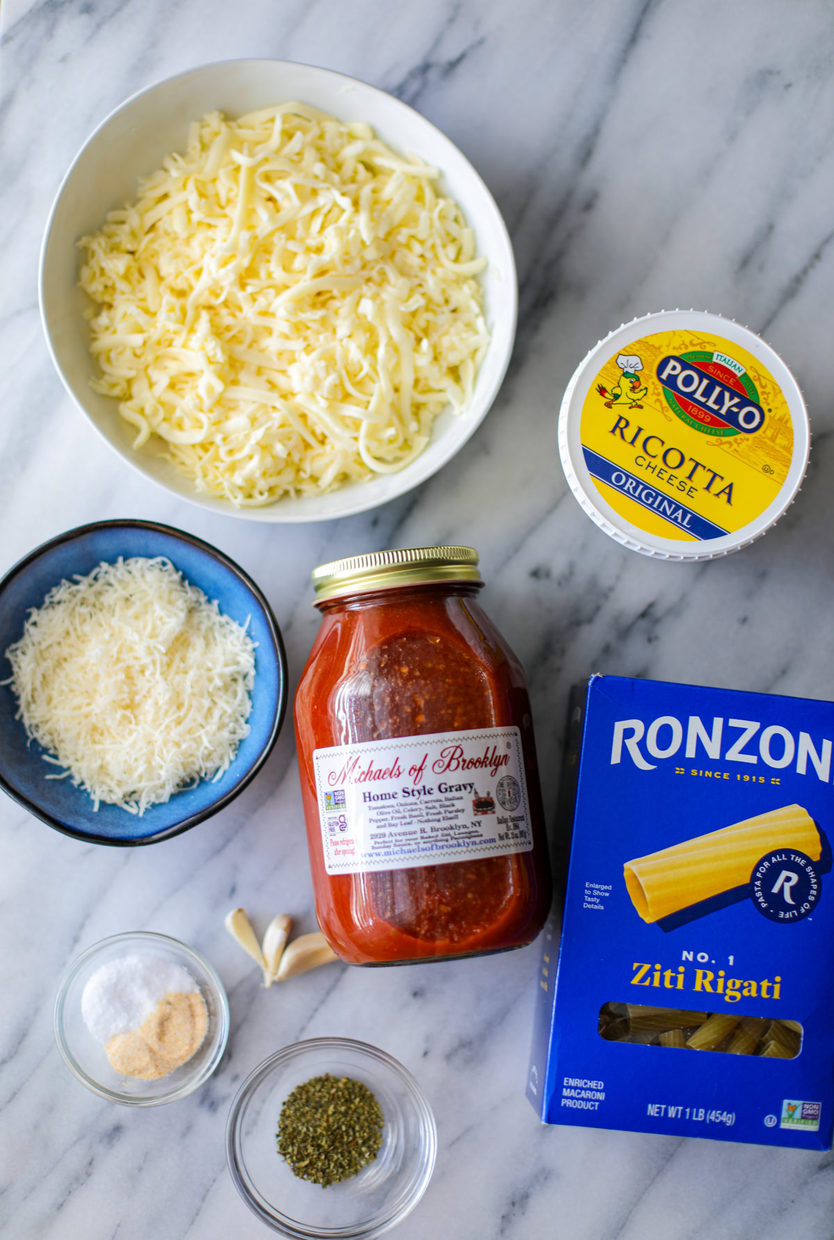 A large white bowl of shredded mozzarella, a yellow container of ricotta cheese, a small blue bowl with shredded parmigiano reggiano, a blue box of Ronzoni Ziti Rigiati pasta, a large mason jar of red home style gravy sauce, two small clear bowls with white salt, onion powder, and green oregano, and 3 unpeeled garlic cloves. 