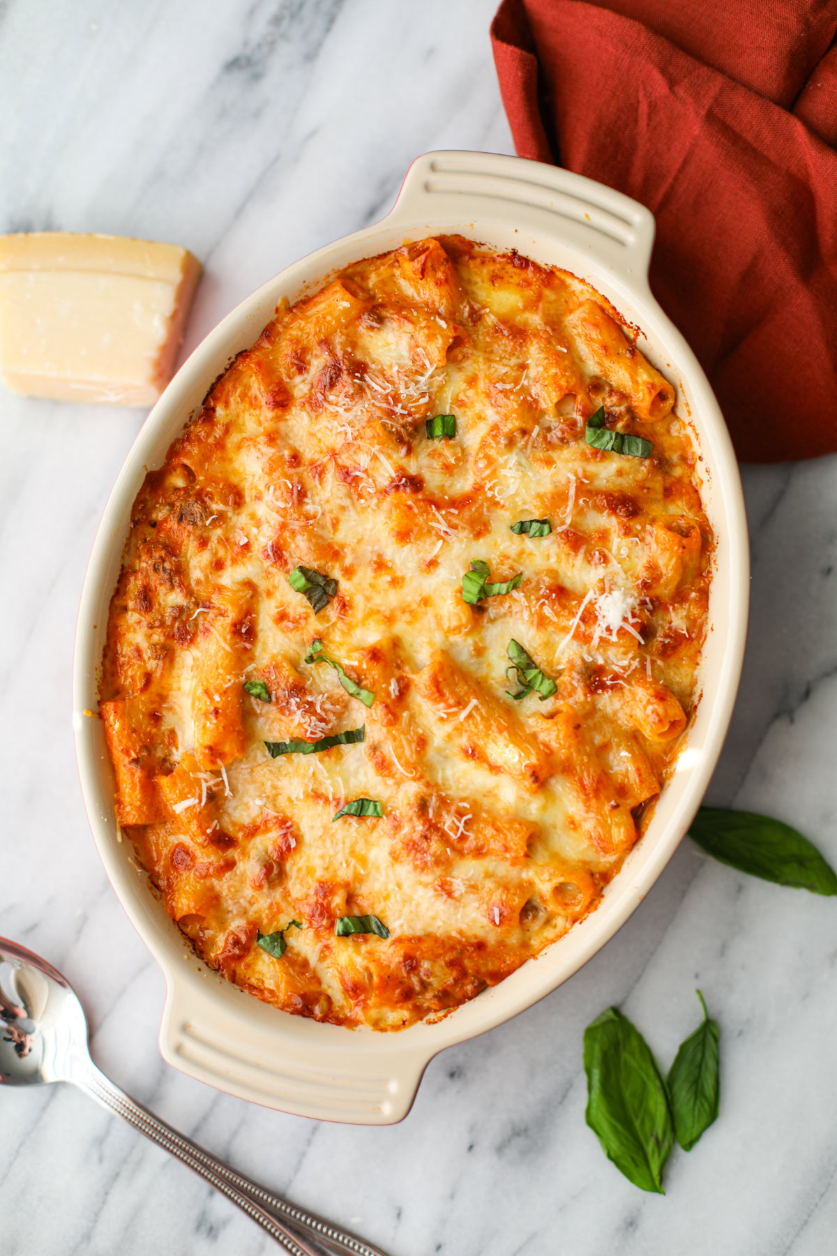 An oblong orange baking dish of baked ziti with crispy, cheese topping and green pieces of fresh basil, with a red napkin, wedge of parmesan cheese, and loose pieces of bright green basil in the background. 