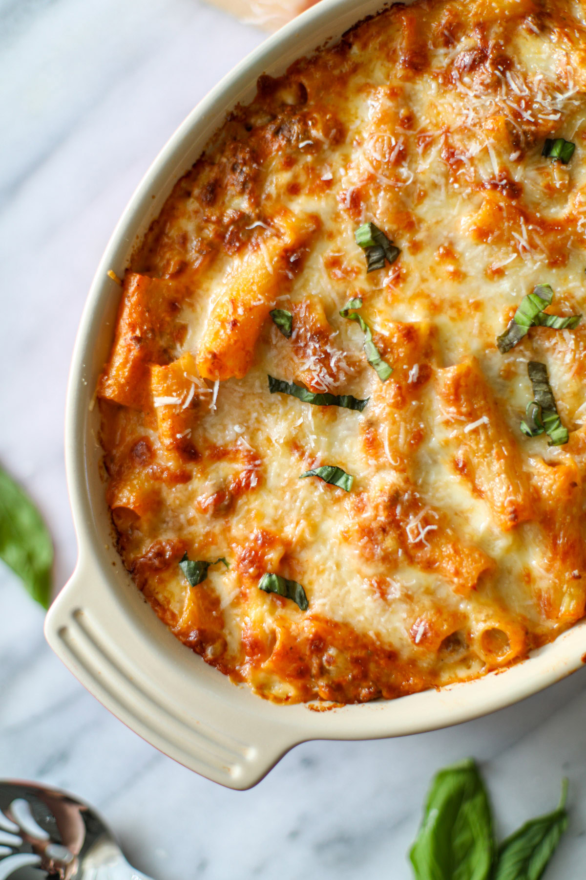 An oblong orange baking dish of baked ziti with crispy, cheese topping and green pieces of fresh basil, with a red napkin, wedge of parmesan cheese, and loose pieces of bright green basil in the background. 