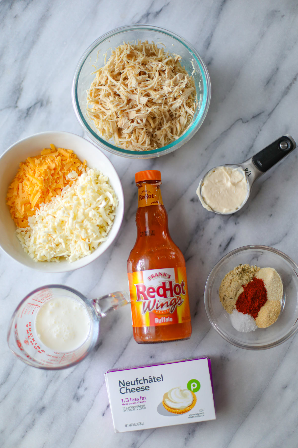 A clear bowl of shredded and cooked chicken, a measuring cup filled with mayonnaise, a bottle of buffalo sauce, a measuring cup with heavy cream, a white bowl half filled with sharp cheddar and half filled with Monterey Jack, a clear bowl of little piles of spices, and a block of Publix brand neufchatel cheese.