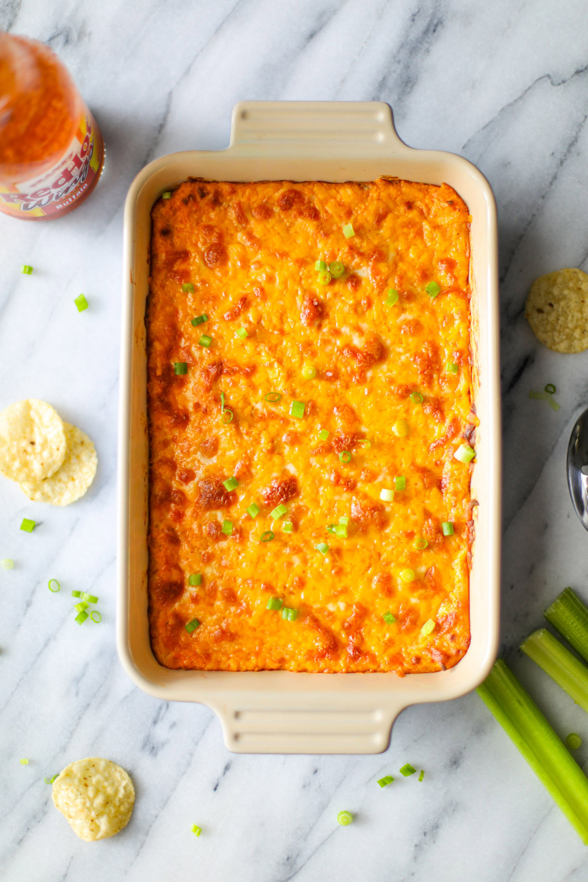 A white and blue oblong baking dish filled with orange and yellow buffalo chicken dip with brown crispy bits and chopped green onions with tortilla chips, sliced celery, chives, and a bottle of franks buffalo sauce surrounding it.