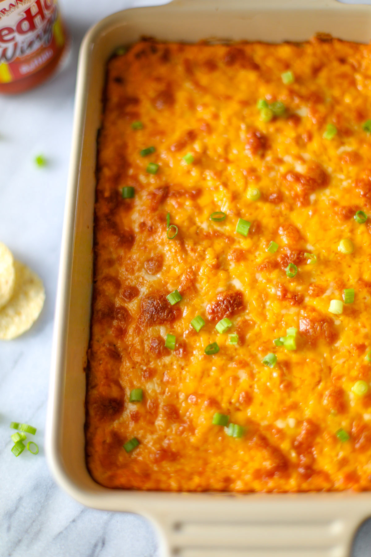 A white and blue oblong baking dish filled with orange and yellow buffalo chicken dip with brown crispy bits and chopped green onions with tortilla chips, sliced celery, chives, and a bottle of franks buffalo sauce surrounding it.