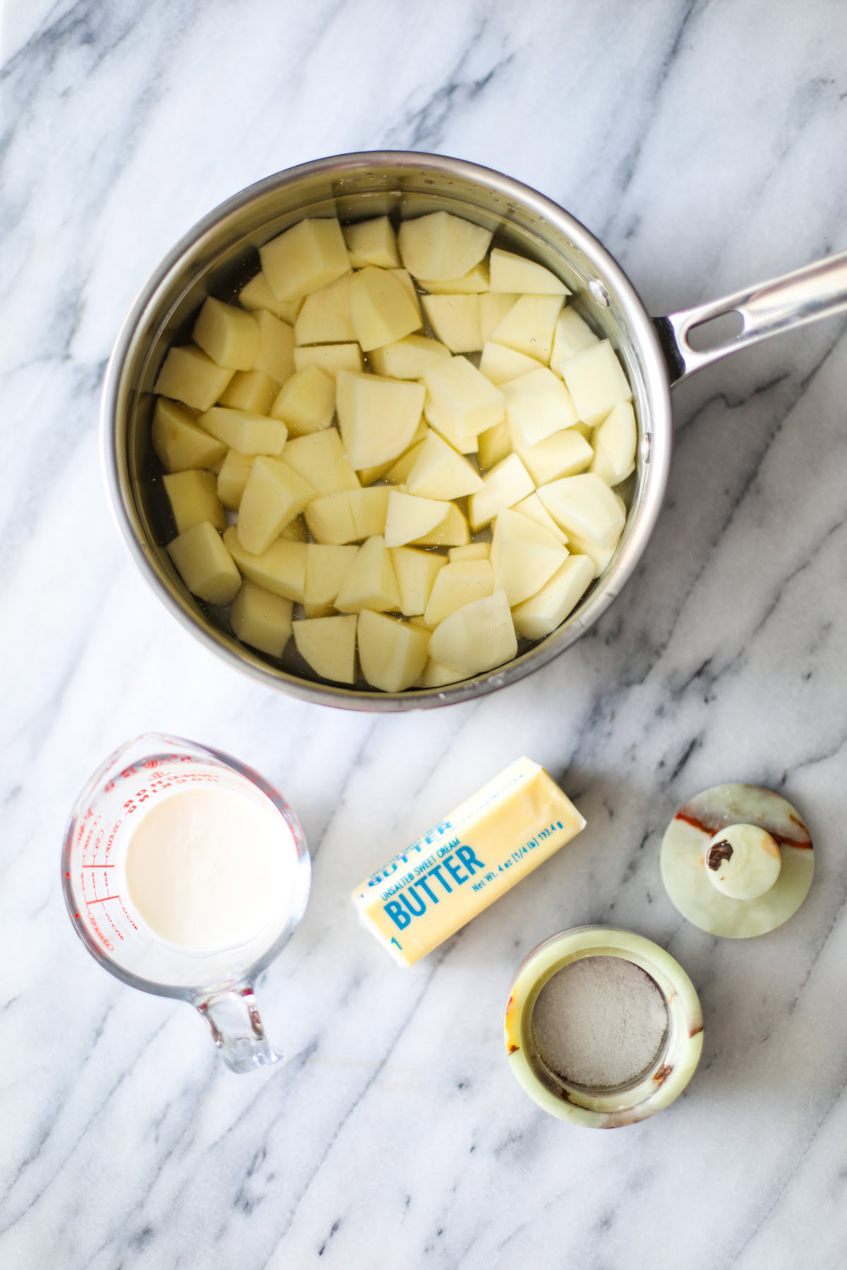 A stainless steel sauce pan filled with chopped and peeled potatoes covered in water, a glass measuring cup filled with white heavy cream, half a stick of butter still in its packaging, a green marble salt cellar filled with salt with it's lid off to the side. 