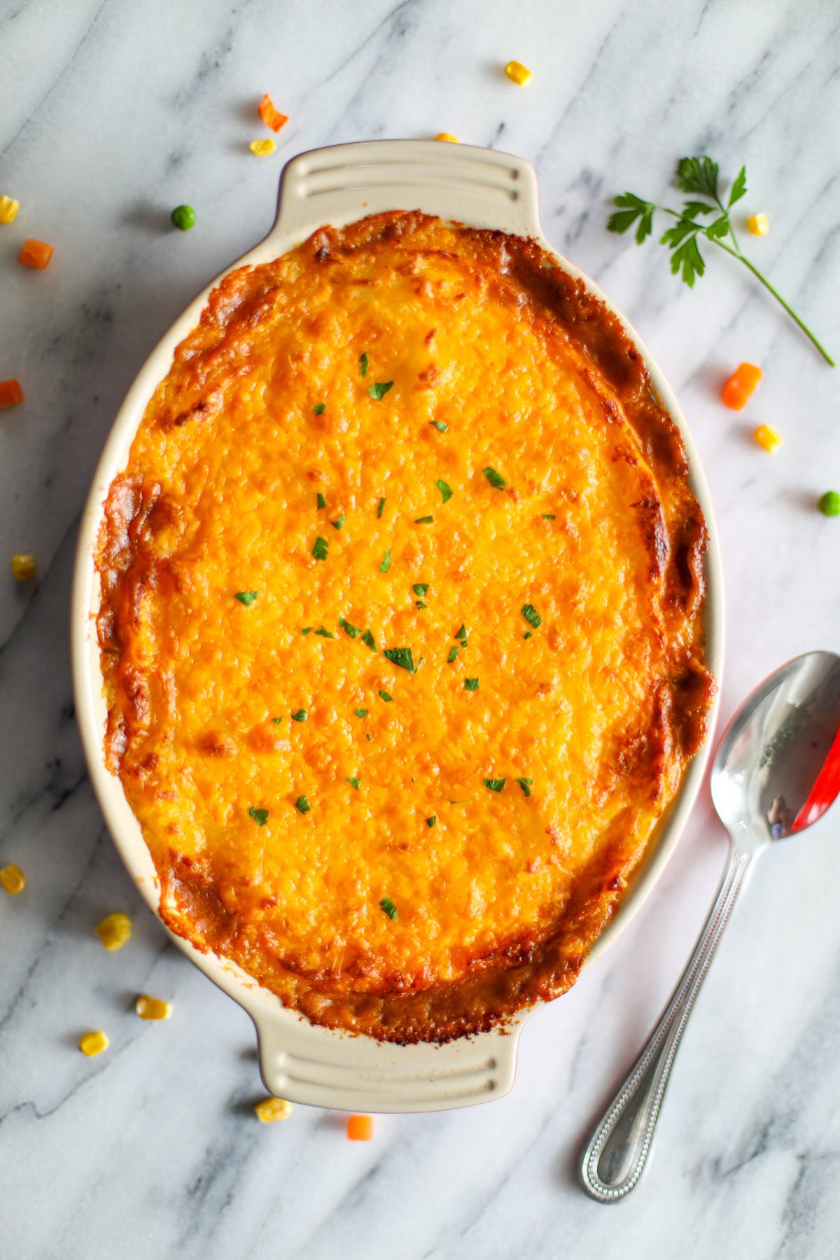 A round oblong casserole dish filled with Cottage Pie with a crispy, cheesy crust and small pieces of chopped parsley to top it with a large metal spoon and loose mixed vegetables in the background.
