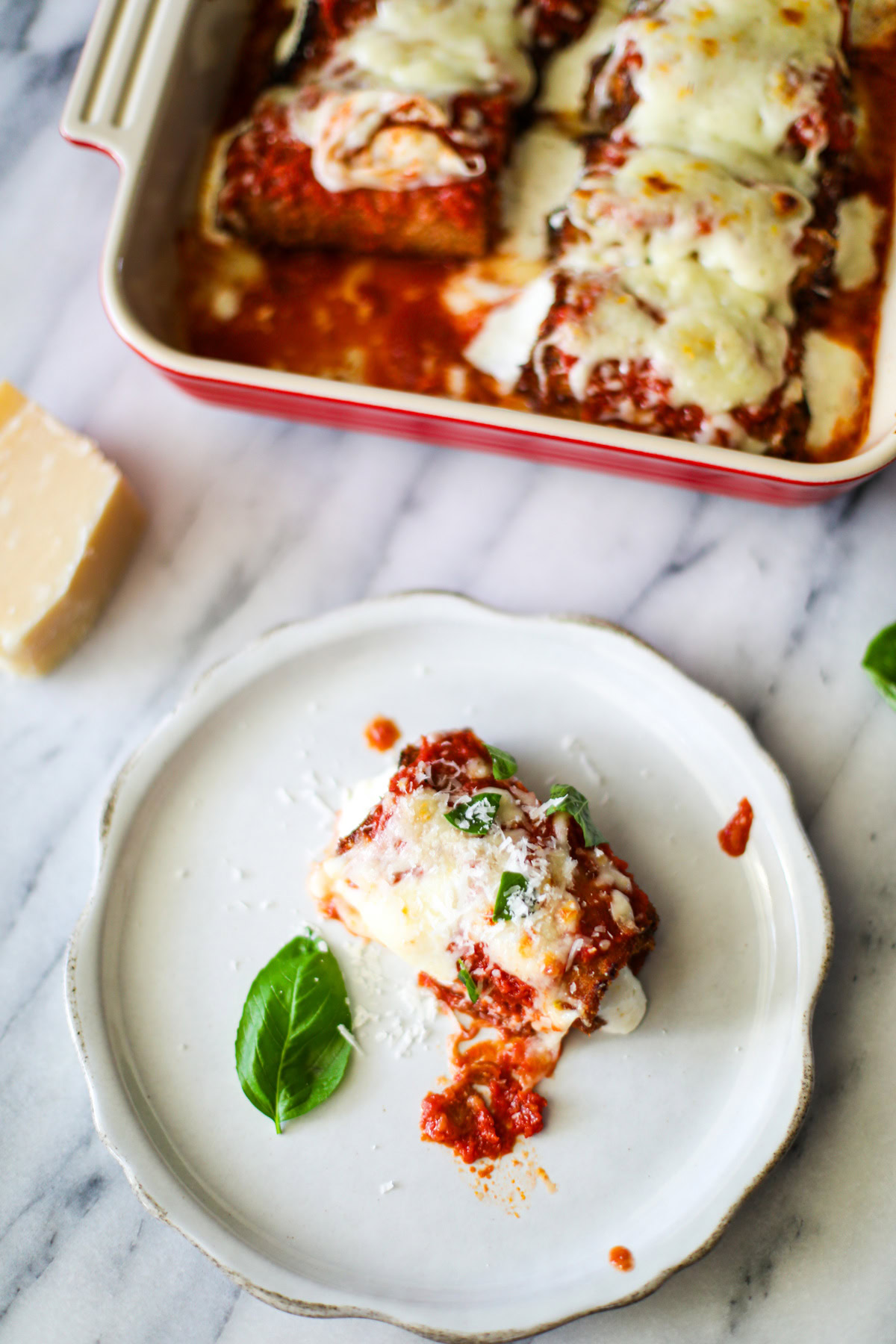 A rustic white plate topped with a rolled up fried piece of eggplant topped with rich red marinara sauce, golden mozzeralla cheese, and pieces of bright green basil with the red baking dish full of rolls sitting in the background.