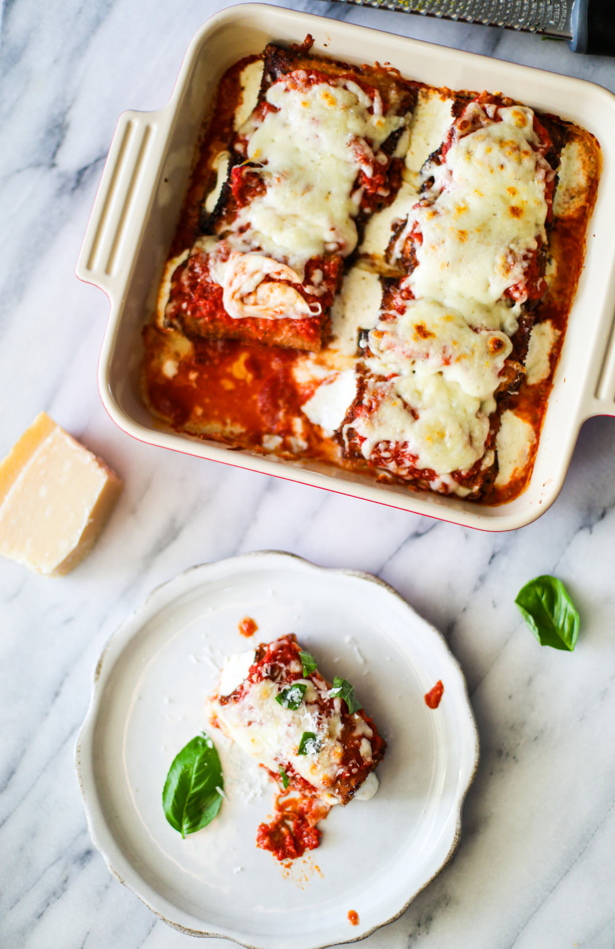 A far away photograph of a rustic white plate topped with a rolled up fried piece of eggplant topped with rich red marinara sauce, golden mozzeralla cheese, and pieces of bright green basil with the red baking dish full of eggplant rollatini sitting in the background.