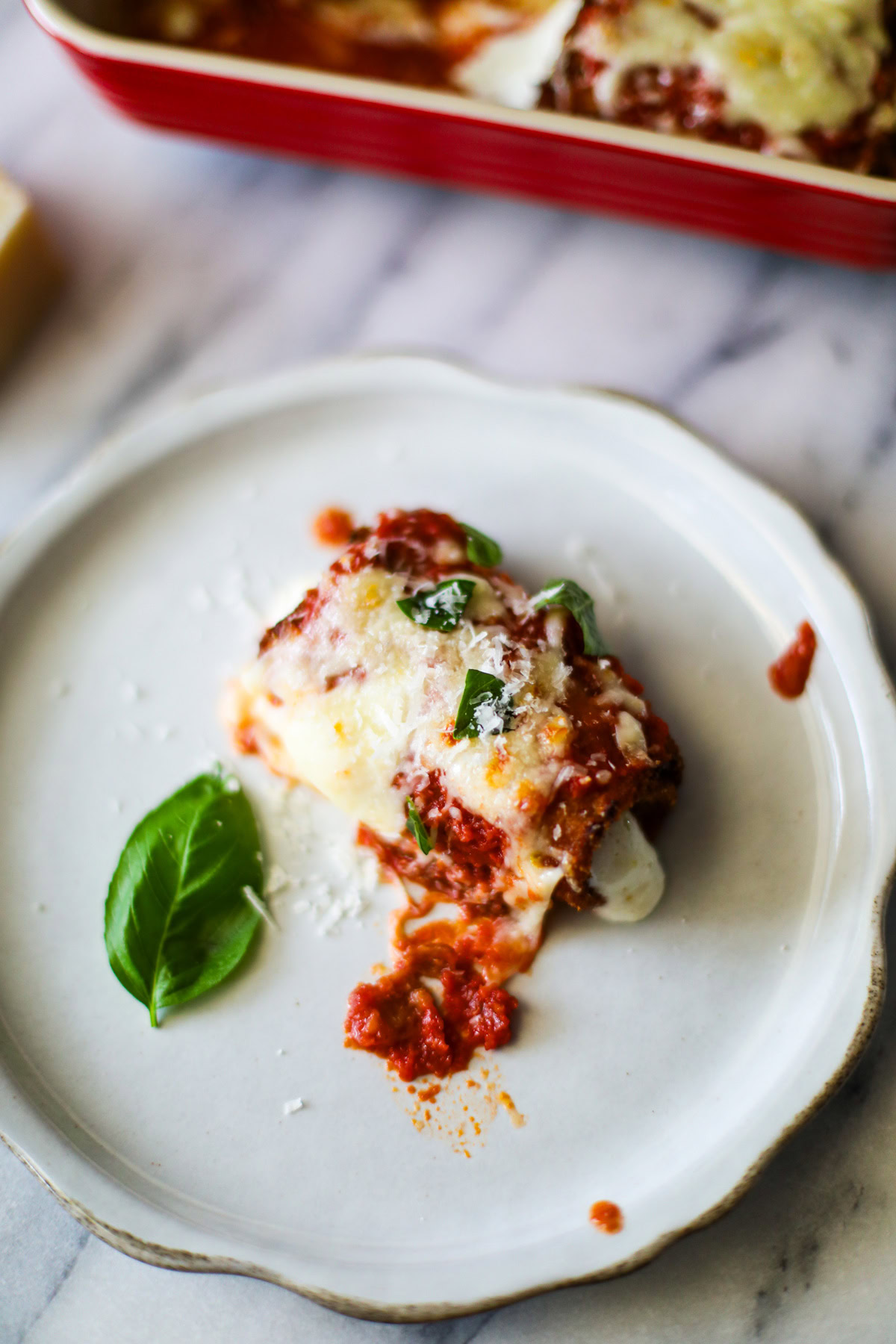 A rustic white plate topped with a rolled up fried piece of eggplant topped with rich red marinara sauce, golden mozzeralla cheese, and pieces of bright green basil with the red baking dish full of eggplant rolls sitting in the background.