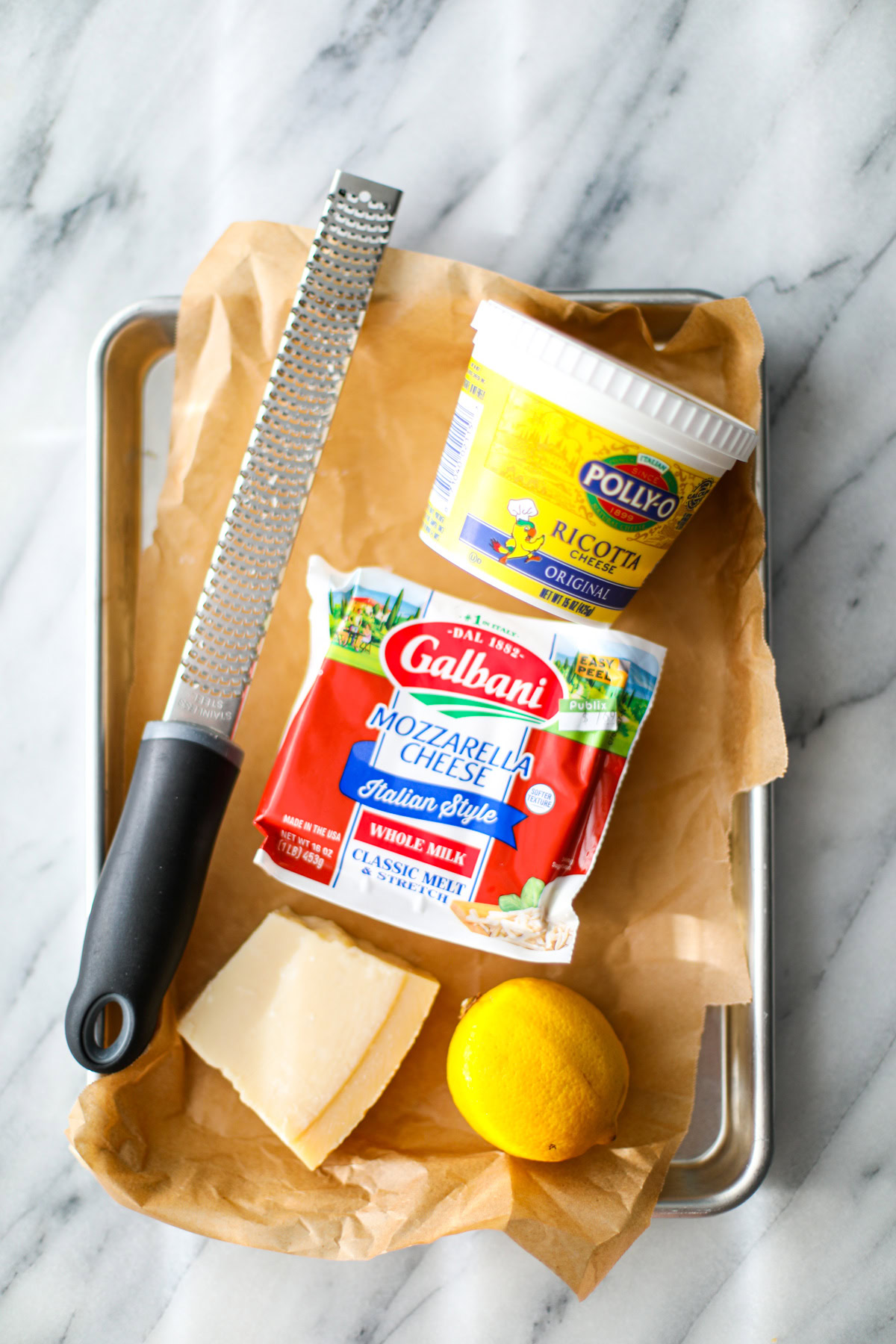 A small sheet pan topped with brown parchment paper with a container of polly-o ricotta, a block of Galvani whole milk mozzarella cheese, a long microplane grater, a whole lemon, and a chunk of parmigiano reggiano to showcase all of the eggplant rollatini filling ingredients.