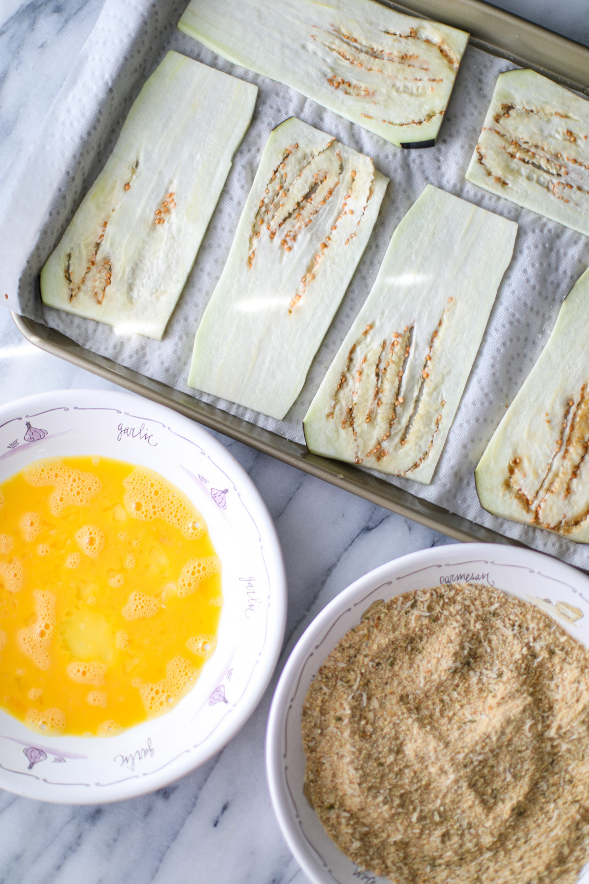 A gold sheet pan with paper towels and slices of eggplant on top, a large shallow bowl of beaten eggs, and another large shallow bowl filled with seasoned Italian breadcrumbs.