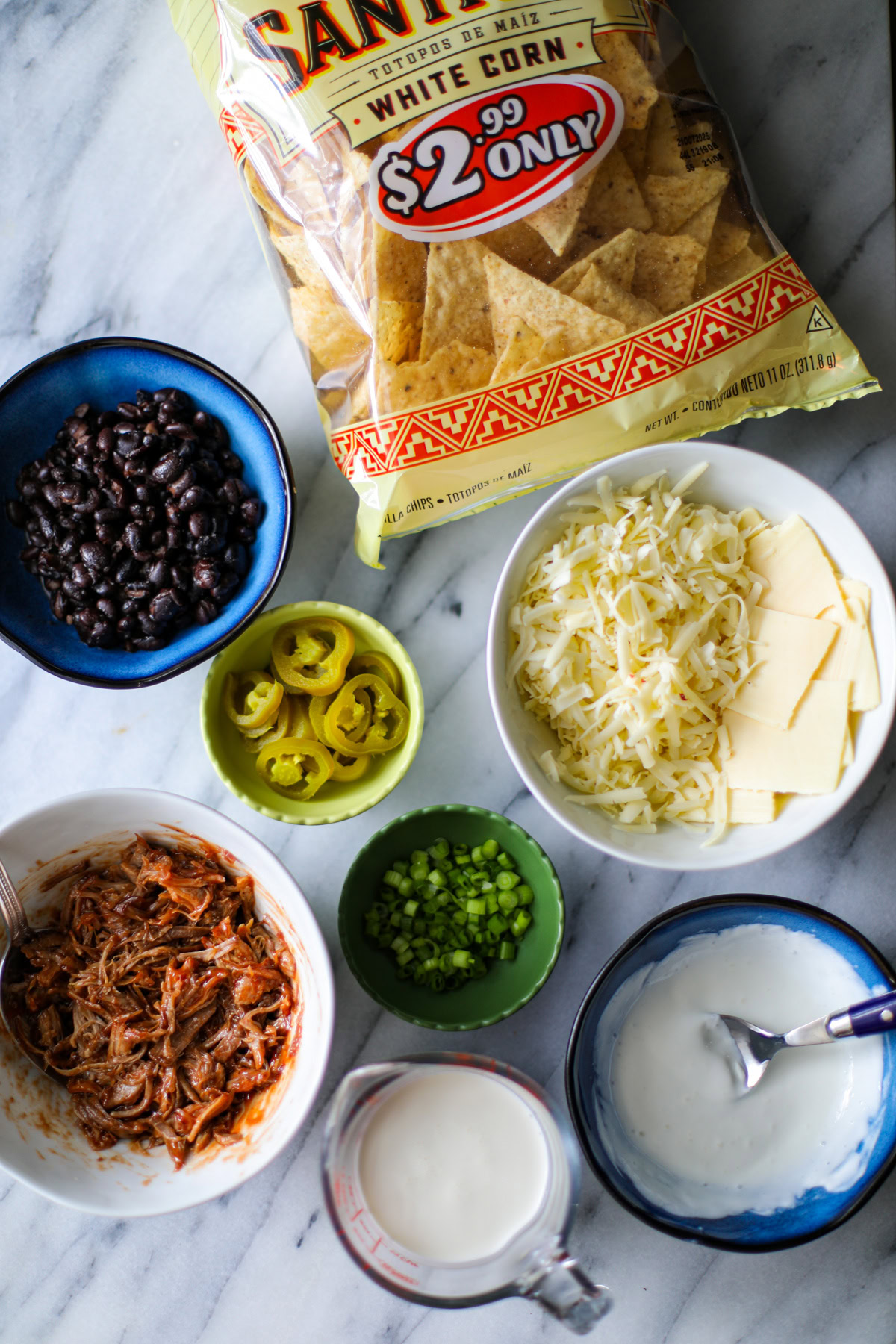 A yellow and red checked bag of white corn tortilla chips, a blue bowl with drained black beans, a white bowl of shredded white cheeses and sliced American cheeses, 2 tiny green pinch bowls with sliced green onions and sliced jalapeños, a white bowl with shredded pulled pork mixed with BBQ sauce, a small blue bowl filled with sour cream with a blue spoon hanging ogg the edge, and a pyrex measuring cup with heavy cream.