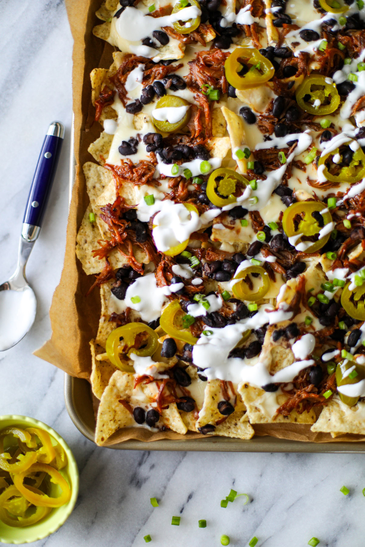 A gold sheet pan lined with brown parchment paper topped with white corn tortilla chips, black beans and brown pulled pork dotted around, with a drizzle of sour cream, a few tamed and sliced jalapeños and thinly sliced green onions scattered over the top. 
