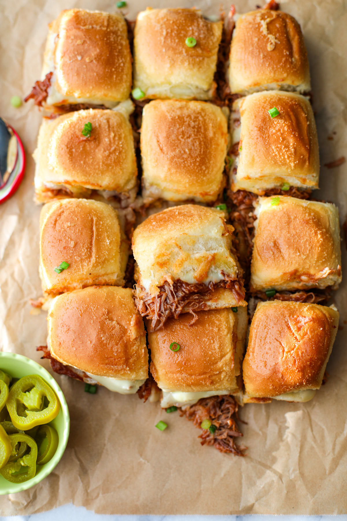 Pulled pork sliders with melted cheese on Hawaiian rolls, topped with green onions, served with pickled jalapeños in a small green bowl sat on top of a brown sheet of parchment paper – perfect party appetizer or game day food.
