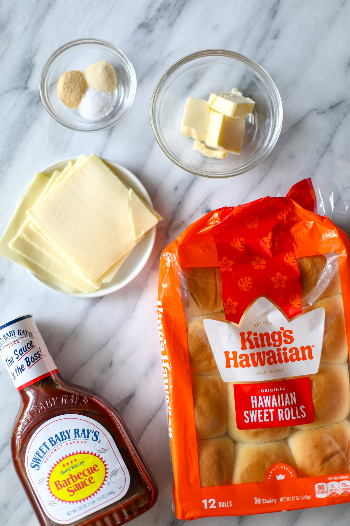 A 12 pack of King's Hawaiian Rolls, a bottle of Sweet Baby Ray's Barbecue Sauce, a small plate of slices of white American cheese, a small clear bowl filled with 3 tablespoons of butter, and a smaller glass bowl with little piles of onion powder, garlic, powder and salt to season the tops of the rolls.