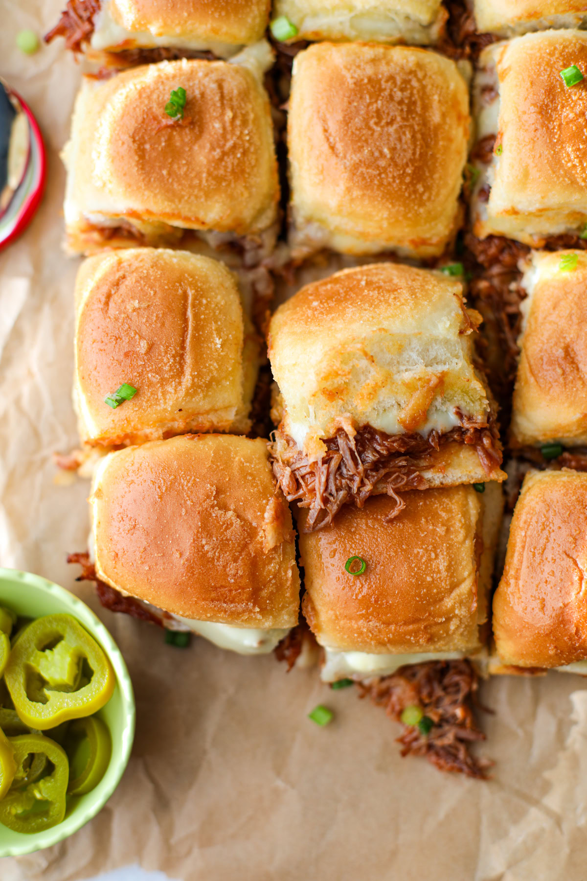 Pulled pork sliders with melted cheese on Hawaiian rolls, topped with green onions, served with pickled jalapeños in a small green bowl sat on top of a brown sheet of parchment paper – perfect party appetizer or game day food.