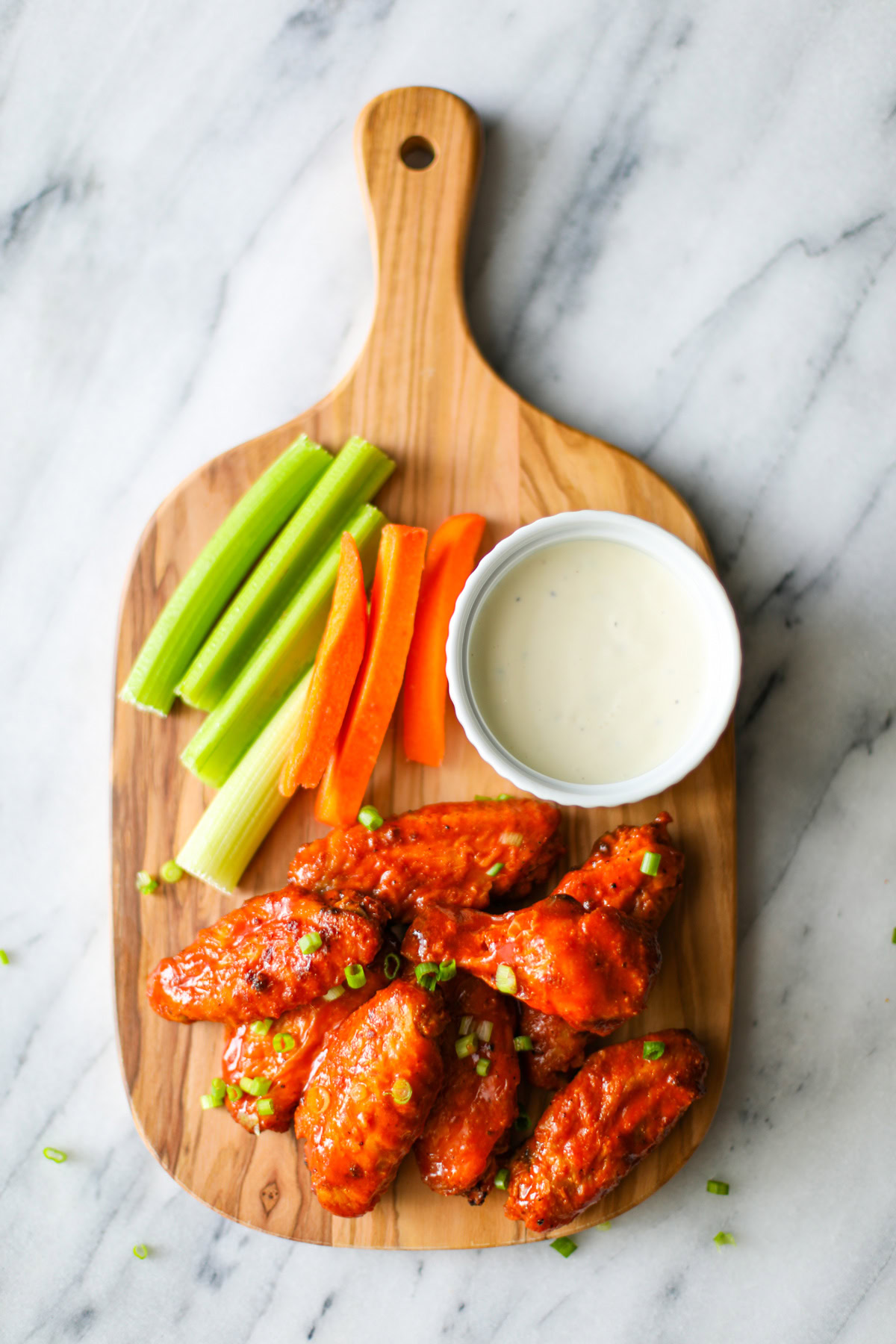 An oak colored serving board sitting on a marble countertop with 7 buffalo wings topped with chopped chives with a white bowl filled with blue cheese and several sticks of carrots and celery placed along the side. 