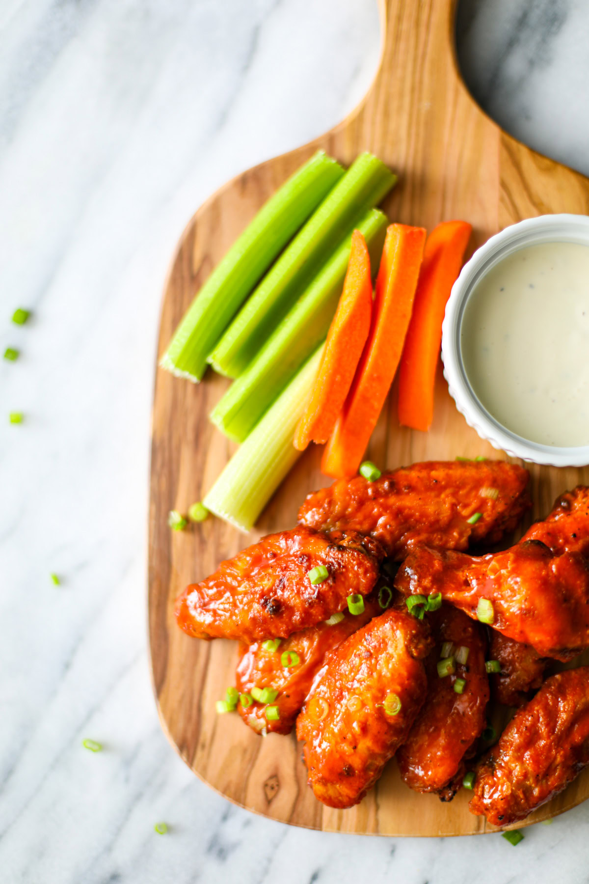 An oak colored serving board sitting on a marble countertop with 7 buffalo wings topped with chopped chives with a white bowl filled with blue cheese and several sticks of carrots and celery placed along the side. 