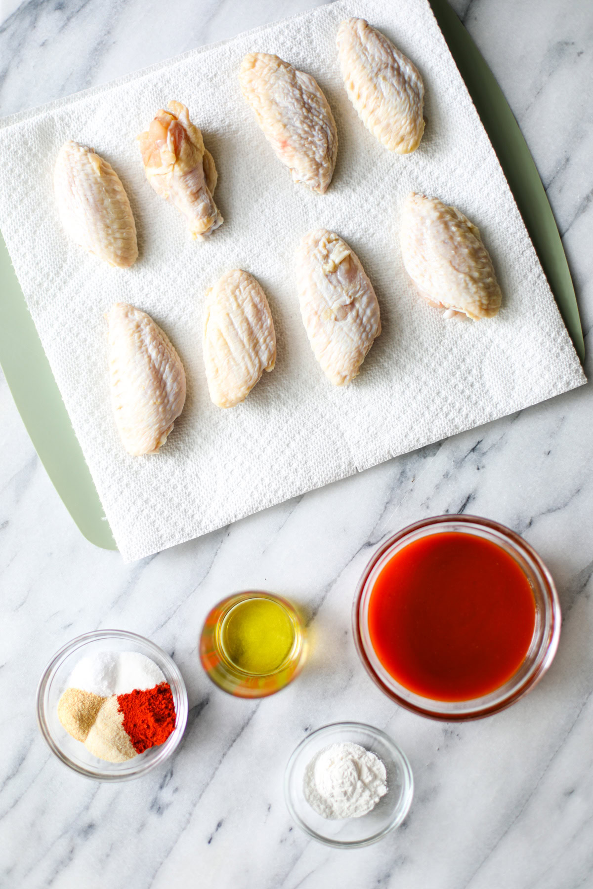 A green cutting board lined with white paper towels with 8 raw chicken wings resting on top, with a small glass bowl of buffalo sauce, a smaller glass bowl of baking powder, and a glass bowl with multiple white and tan seasonings, and a cylindrical measuring cup with olive oil. 