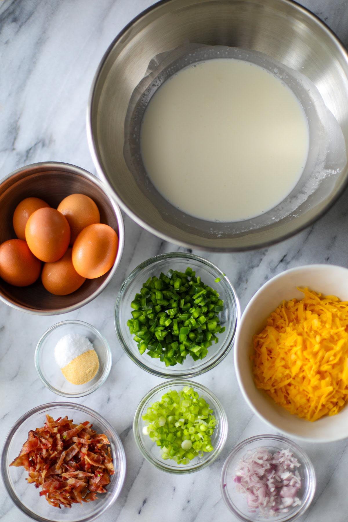 All of the ingredients to make jalapeño popper quiche including a bowl of milk, eggs, jalapeños chopped, shredded cheese, seasonings, bacon pieces, shallot, and sliced green onions.