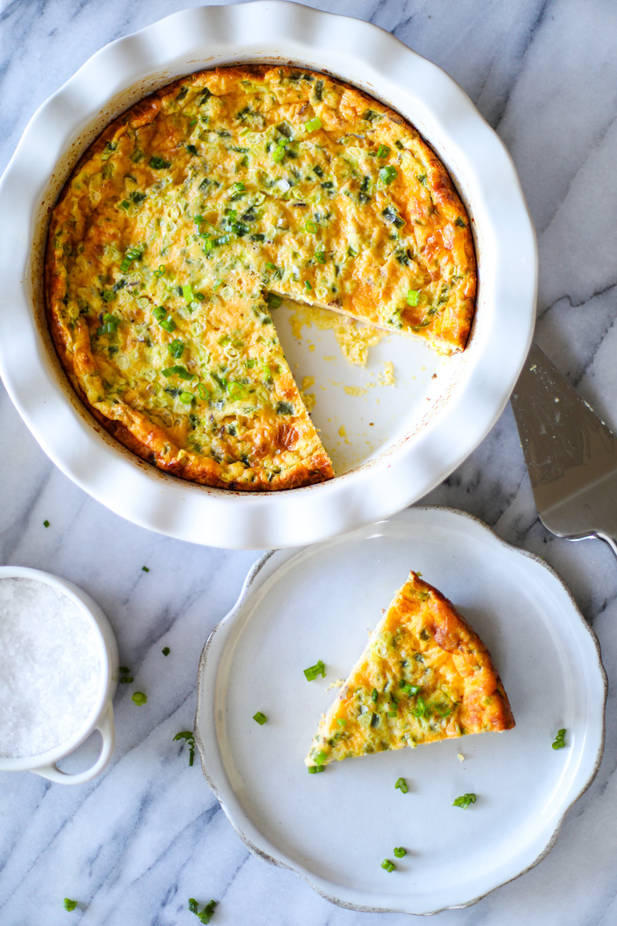 A white ruffled pie dish with jalapeño popper quiche, with a slice on the plate and a pie cutter.