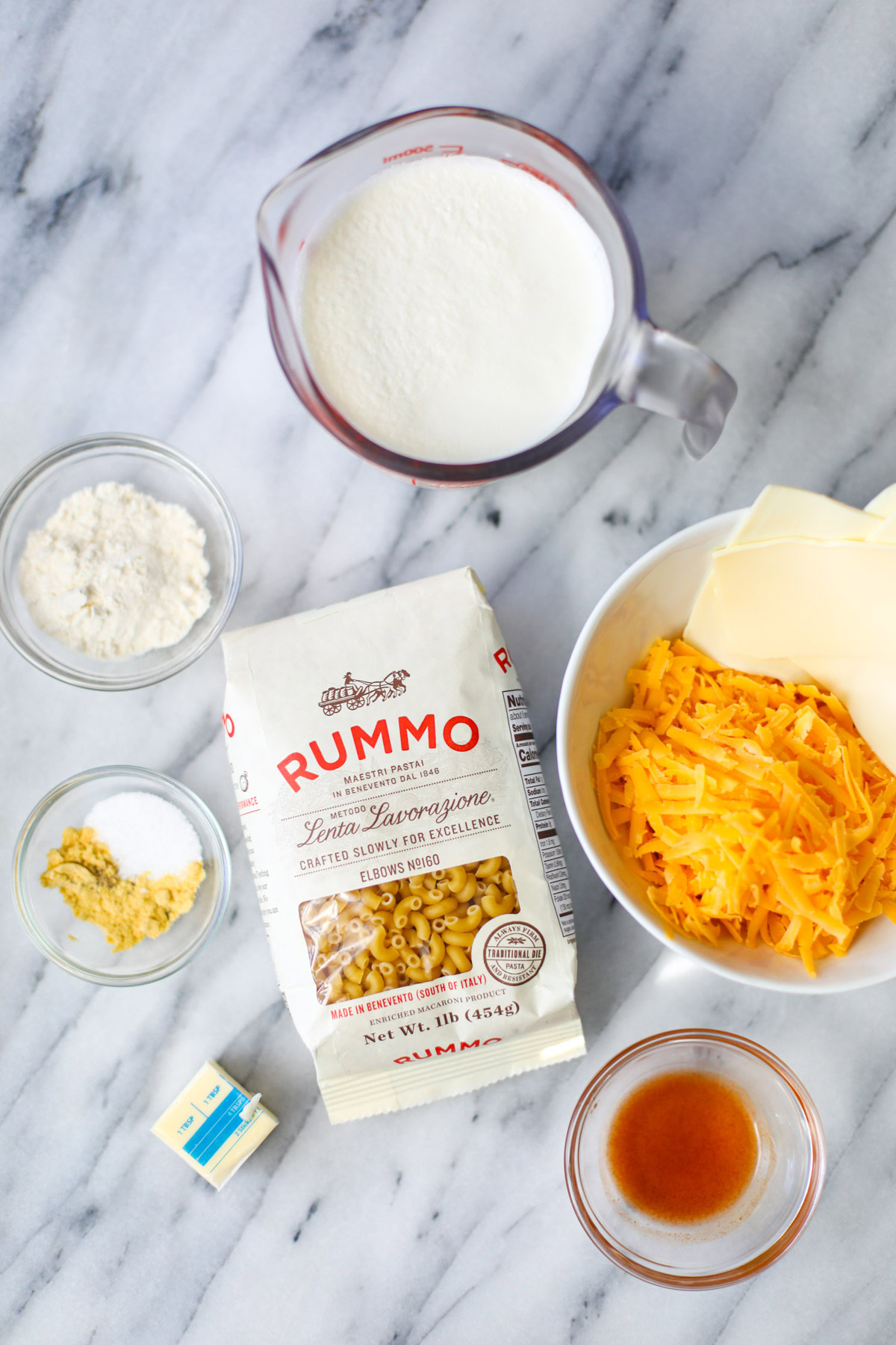 A measuring cup of heavy cream and milk, a bowl of shredded cheese, a bag of elbow pasta, a nob of butter, and two small bowls of seasoning and hot sauce.