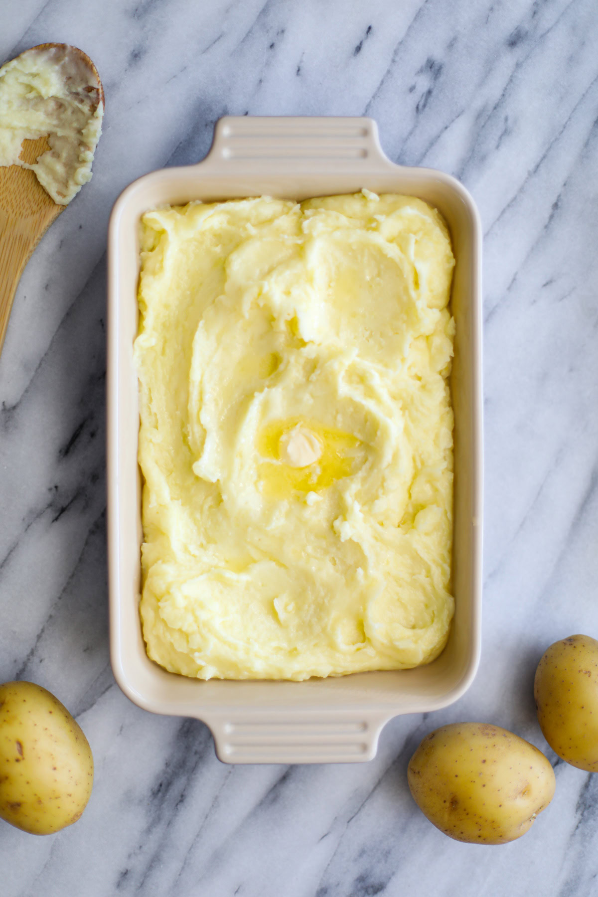A serving dish of mashed potatoes with a nob of melted butter and potatoes surrounding the dish in the background.