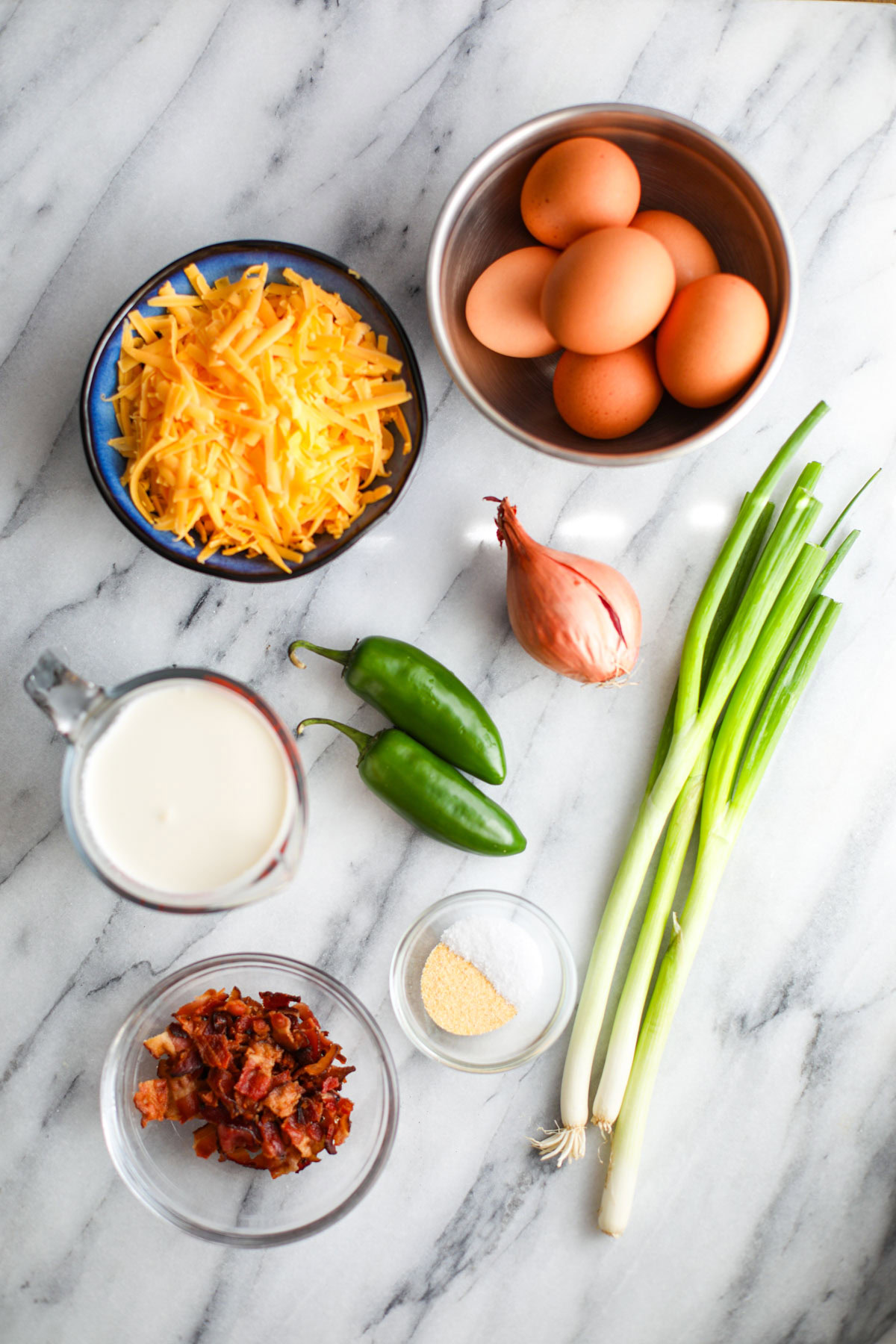 All of the ingredients to make jalapeño popper quiche including a bowl of milk, eggs, jalapeños chopped, shredded cheese, seasonings, bacon pieces, shallot, and sliced green onions.
